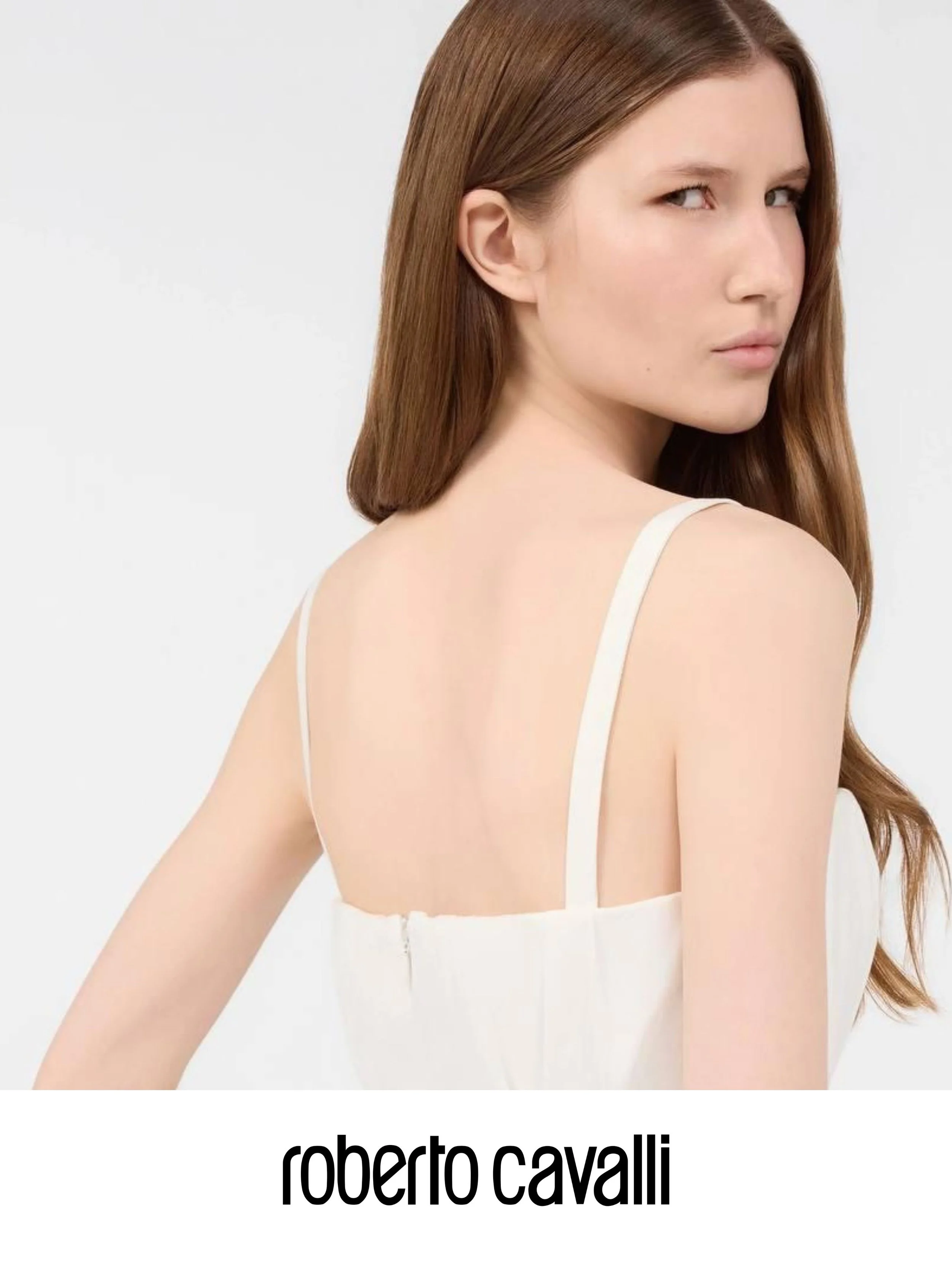 Close-up of a woman with long, straight, brown hair looking over her shoulder against a white background, wearing a white spaghetti strap top, with the text 'roberto cavalli' in black at the bottom.