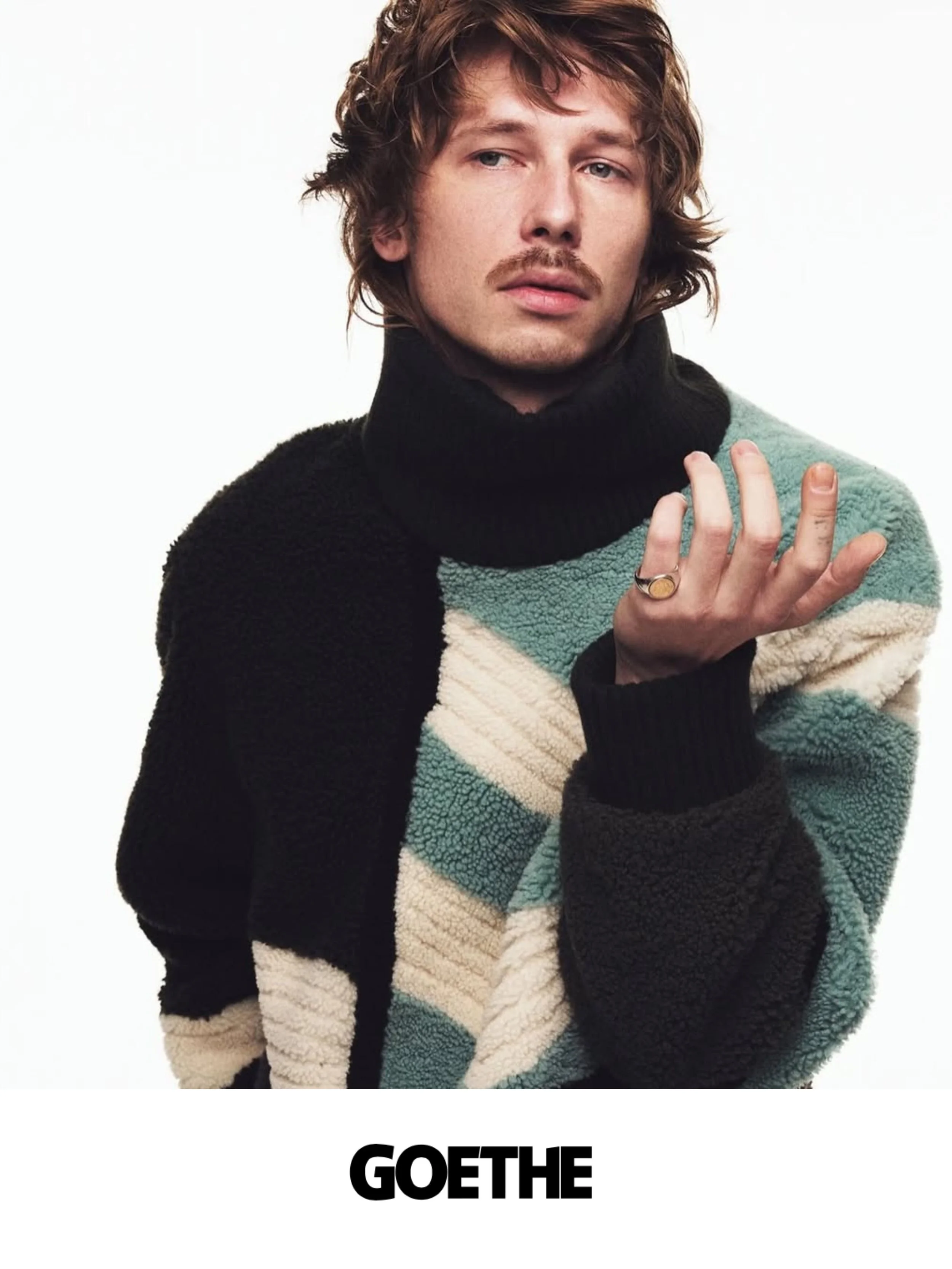 A young man with wavy brown hair and a mustache, wearing a multicolored fuzzy sweater and a ring on his left hand, poses against a plain white background. The word "GOETHE" appears in bold black letters below the image.