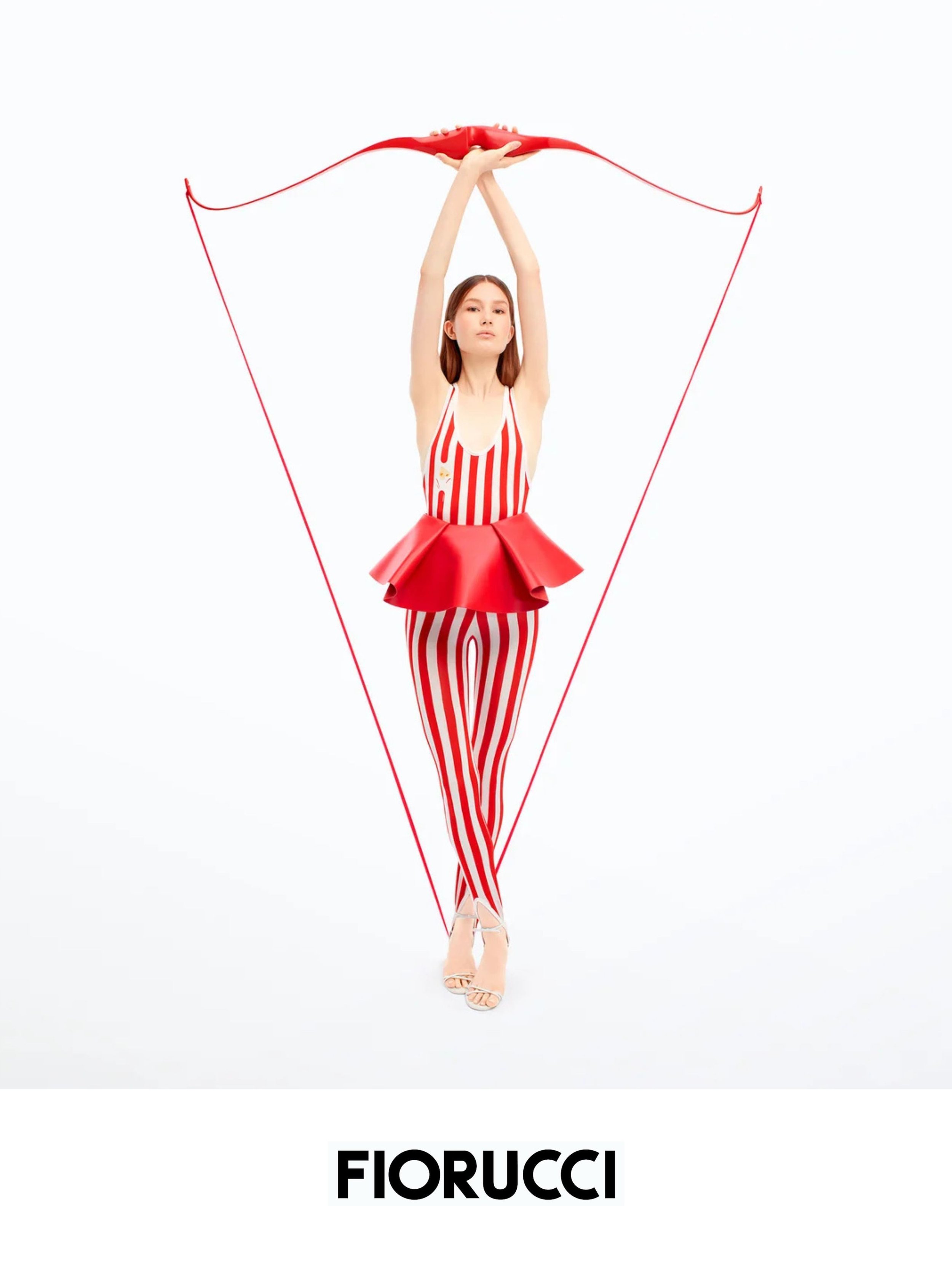 A woman in a red and white striped dress and matching pants stands on heels, holding a red bow above her head with both hands, in a white studio with minimal background. Below her, the word 'FIORUCCI' is displayed in bold black letters.