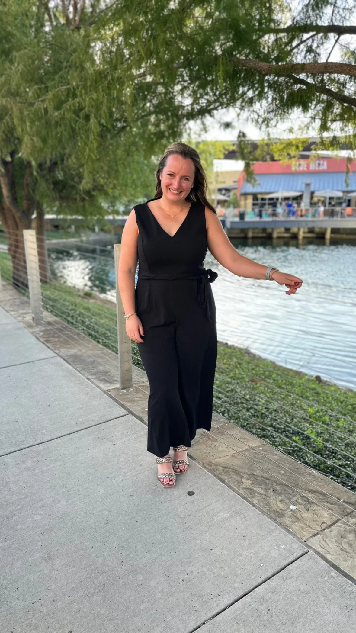 A woman in a black sleeveless jumpsuit and high-heeled sandals standing on a sidewalk near a body of water at Grand Hyatt  Baha Mar Resort