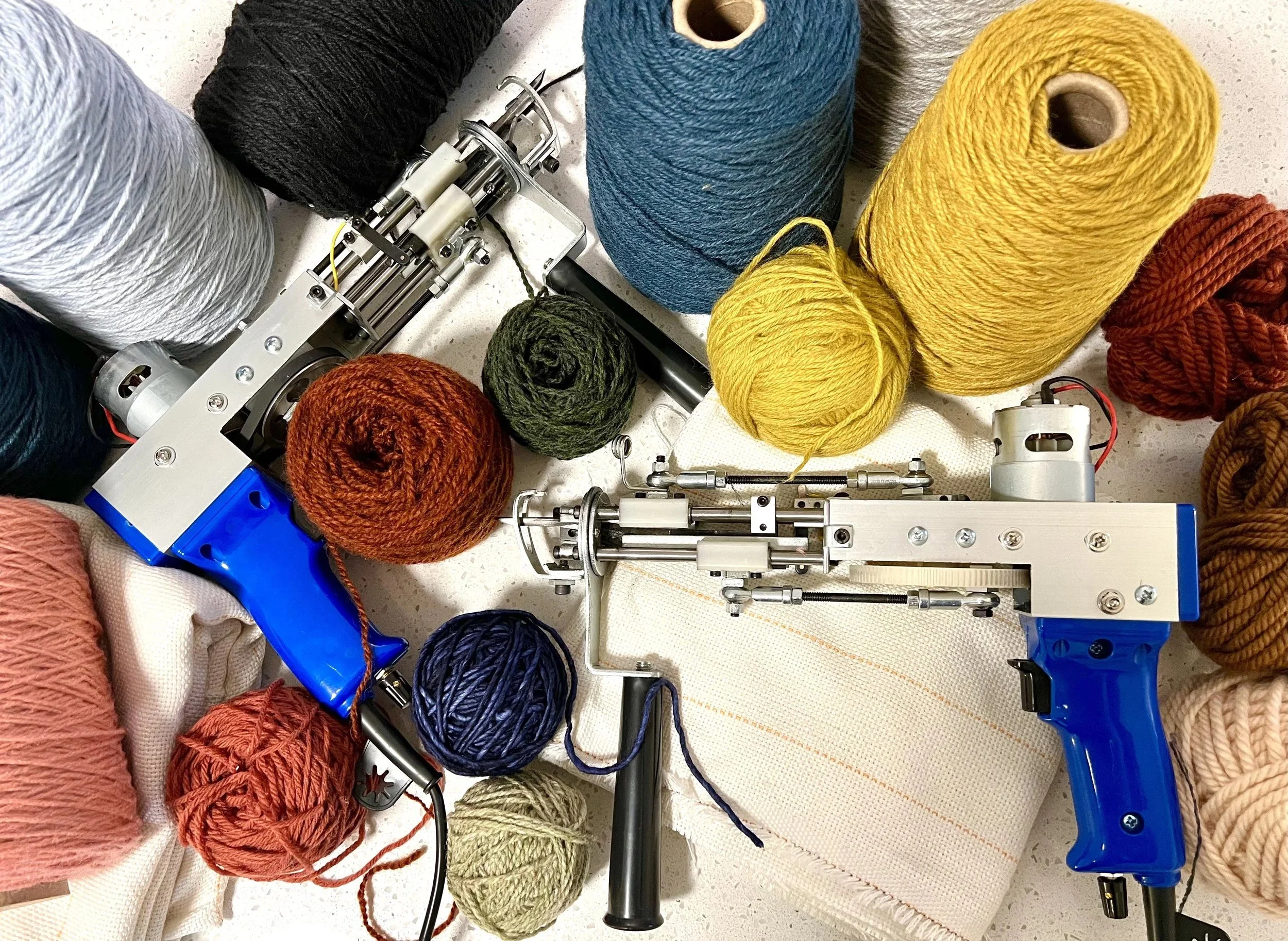 Spindle knitting machine surrounded by balls of multicolored yarn in shades of blue, yellow, orange, red, pink, green, and cream. The machine has metallic parts and a blue and white body, with some yarn threads attached.