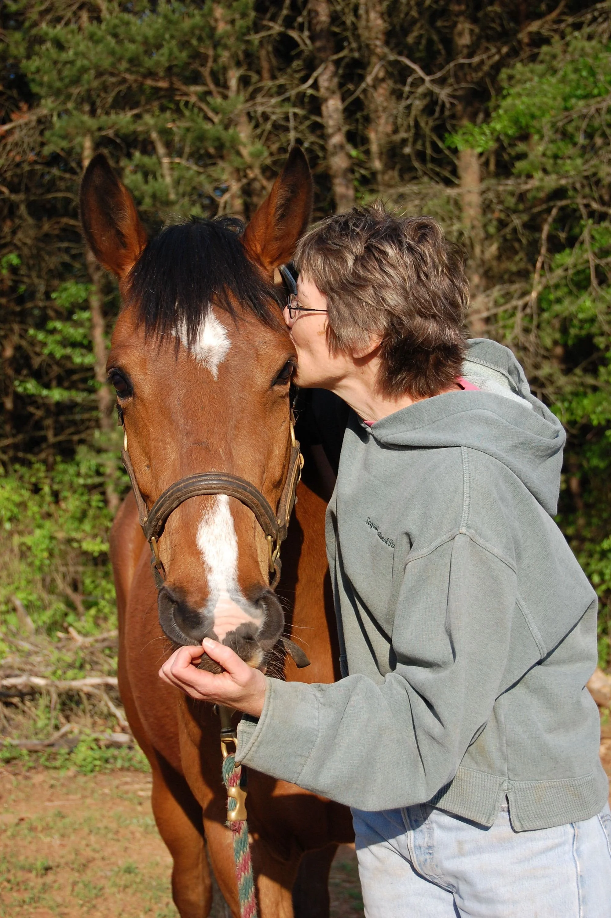 Rett kissing Ayla (the horse) on the face while holding its near nostril with their hand, outdoors with green trees in the background.