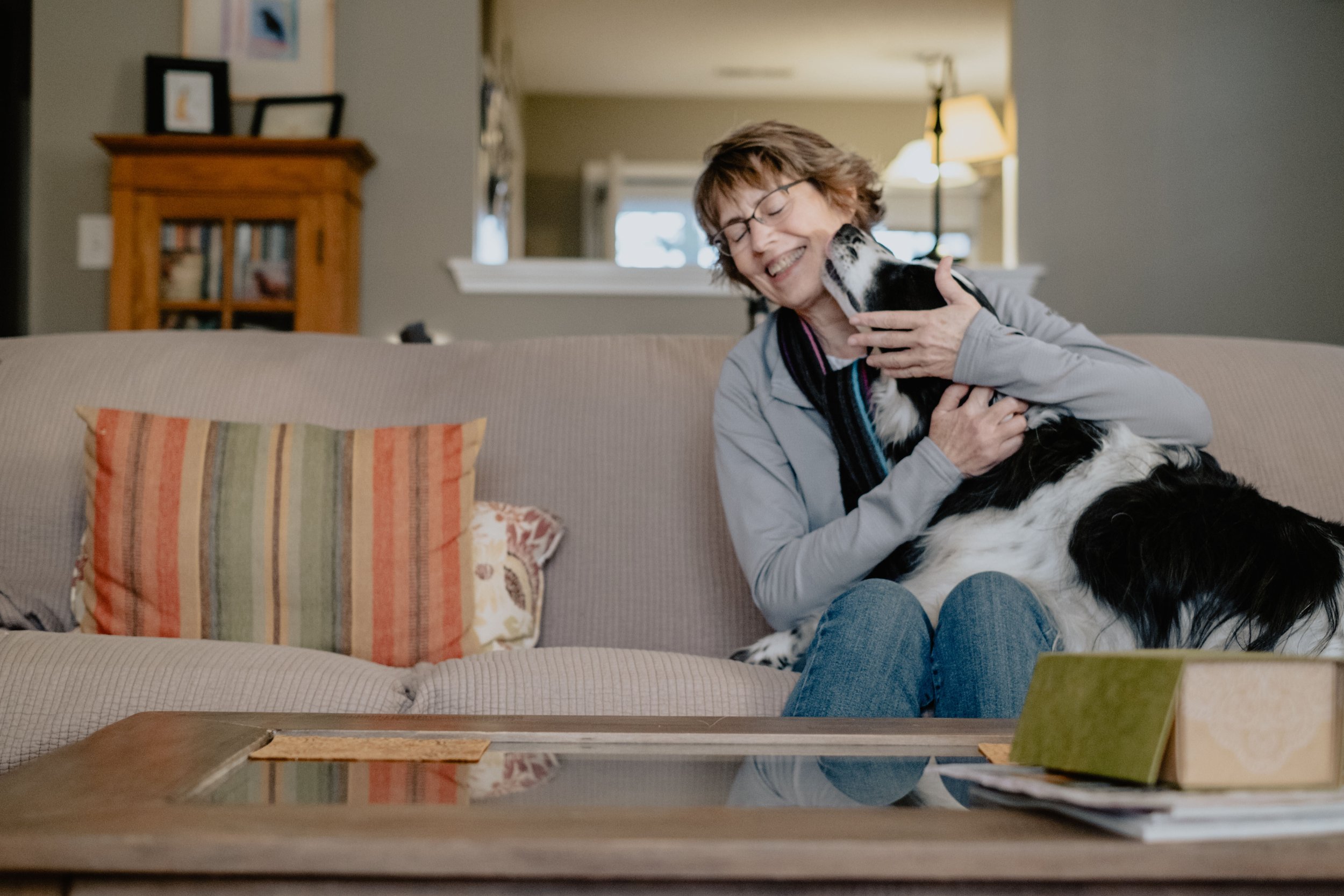 Rett sitting on a beige couch, smiling and holding Piper her dog, in a cozy living room with a wooden bookshelf and a window in the background.