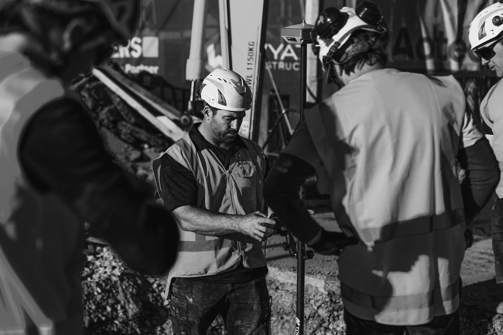 Group of construction workers wearing safety vests and helmets, gathered around a piece of equipment at a worksite.