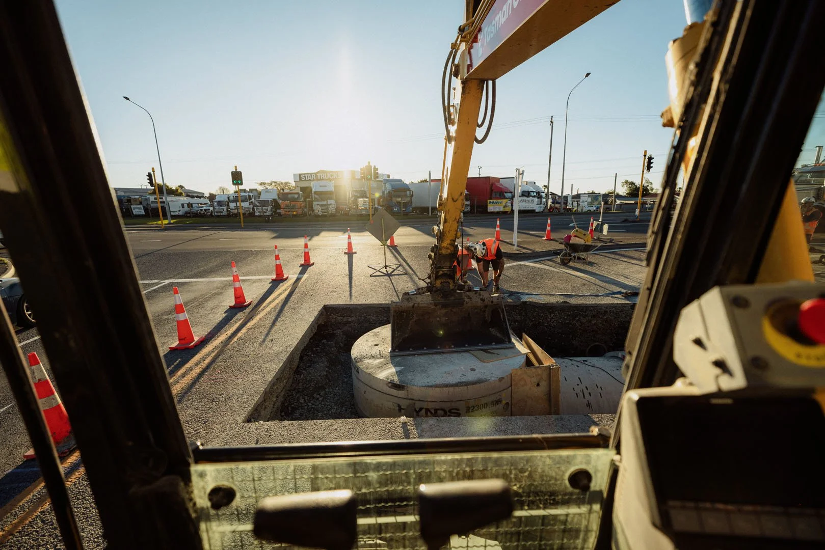 View from inside construction equipment looking out at workers in safety vests and helmets repairing a road, with traffic cones, a busy street, trucks, and the sun setting in the background.