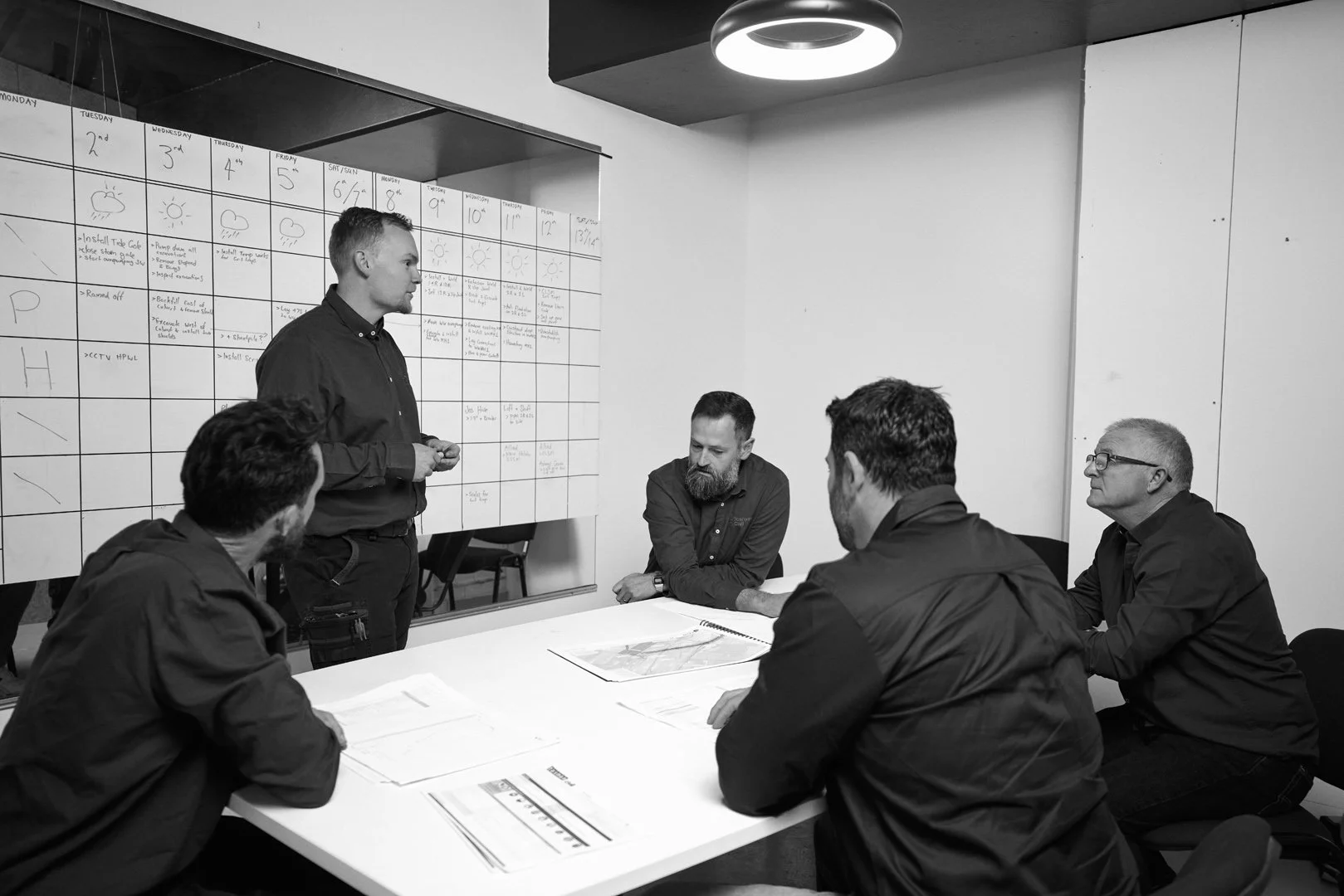 A man is standing and presenting to four seated men in a meeting room. A large whiteboard with a weekly calendar and notes is on the wall behind them.