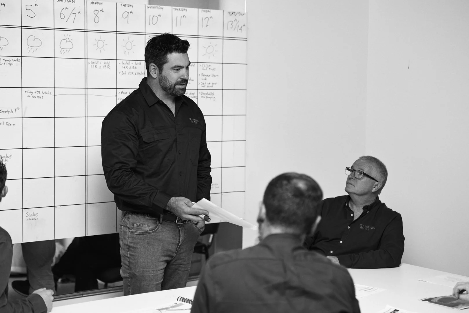 A man is standing and speaking to a group of seated men in a meeting room. The man is holding papers, and a whiteboard with a calendar is visible in the background.