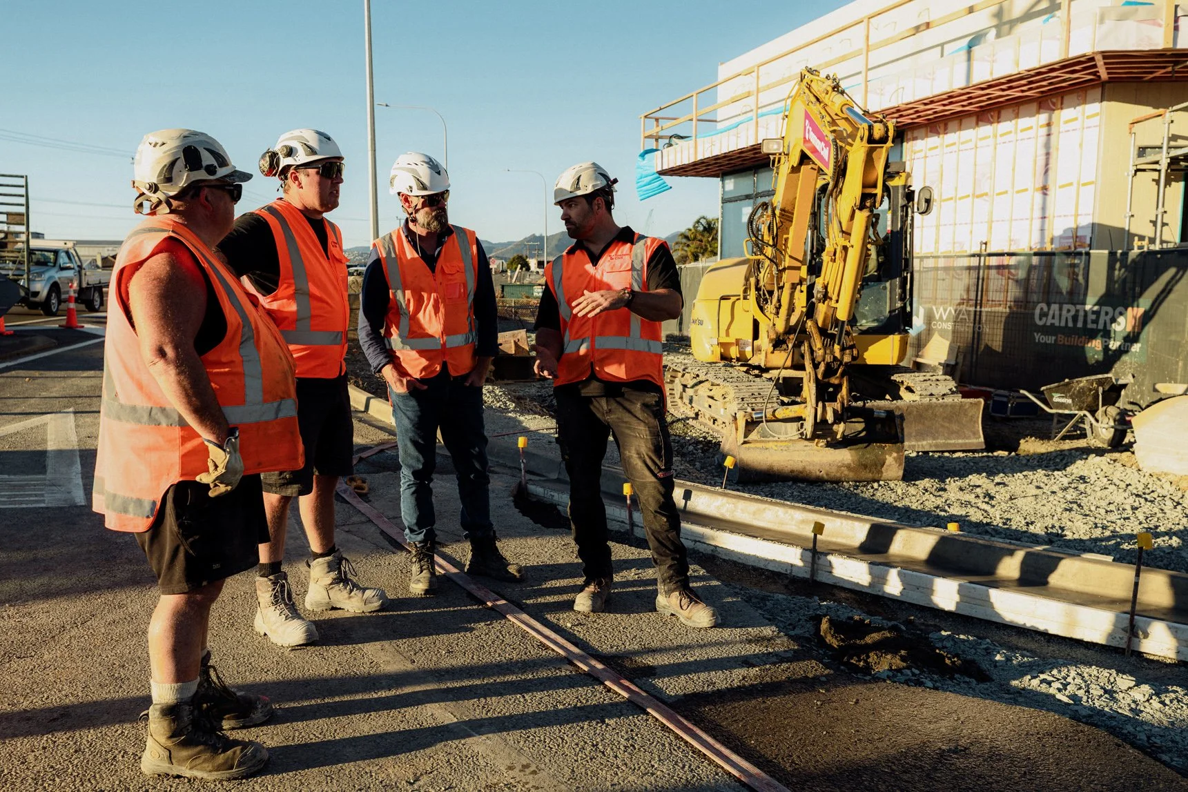 Four construction workers wearing orange safety vests and white helmets stand on a construction site discussing, with a yellow excavator and building materials in the background.