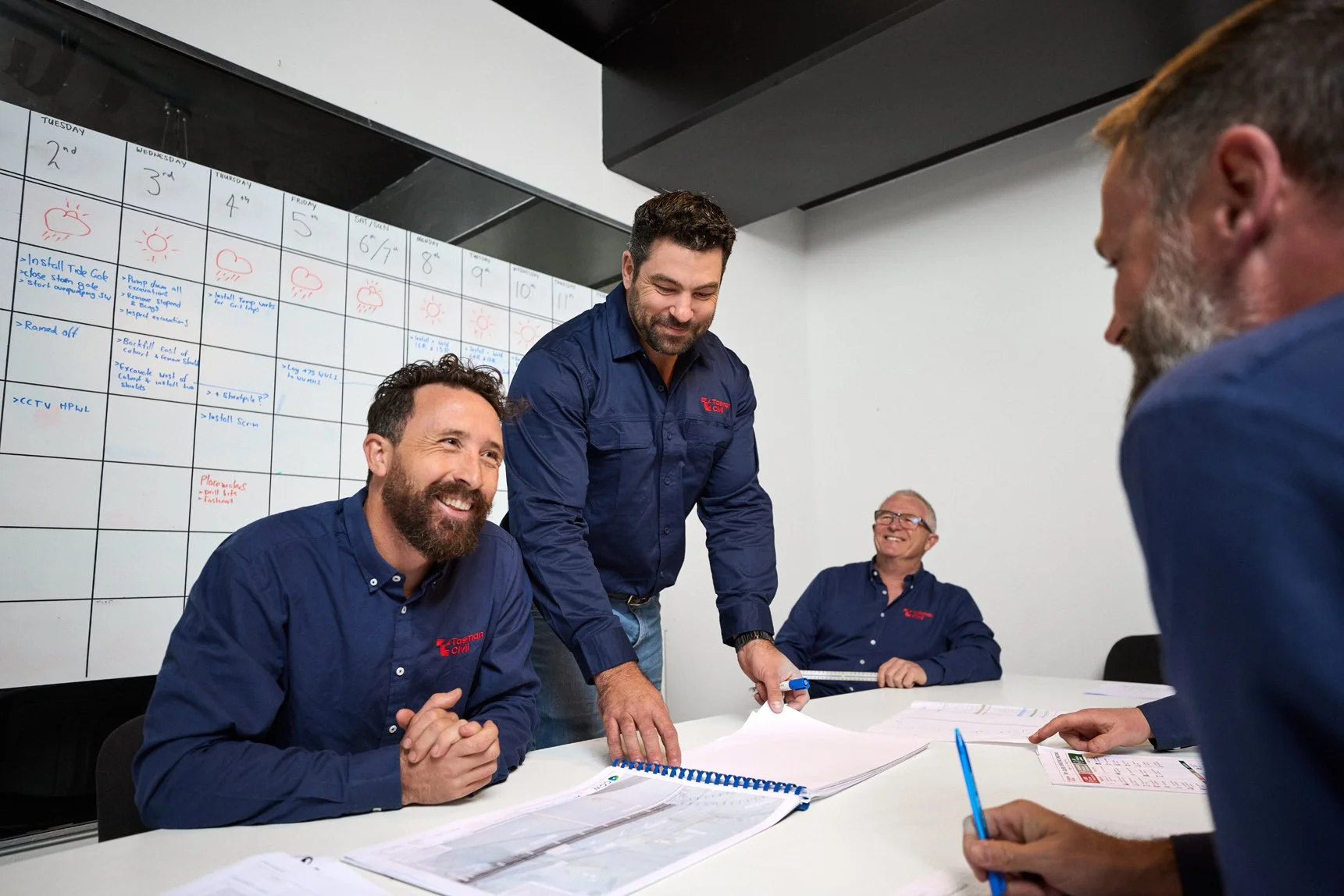 Four men in blue shirts with 'Tasman Civil' logo in a meeting room, one standing and handing documents to seated men, with a whiteboard filled with notes and weather icons in the background, smiling and engaging in discussion.