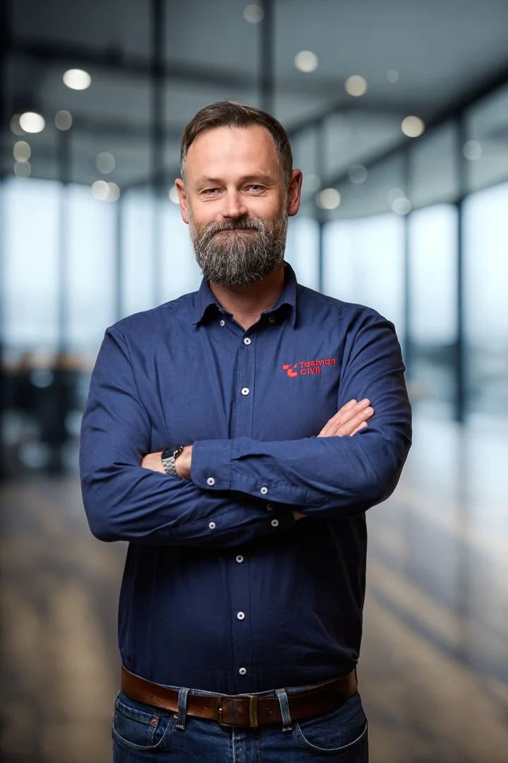 A confident man with a beard, wearing a navy blue shirt with a company logo, inside a modern office building with glass walls and bright lighting.