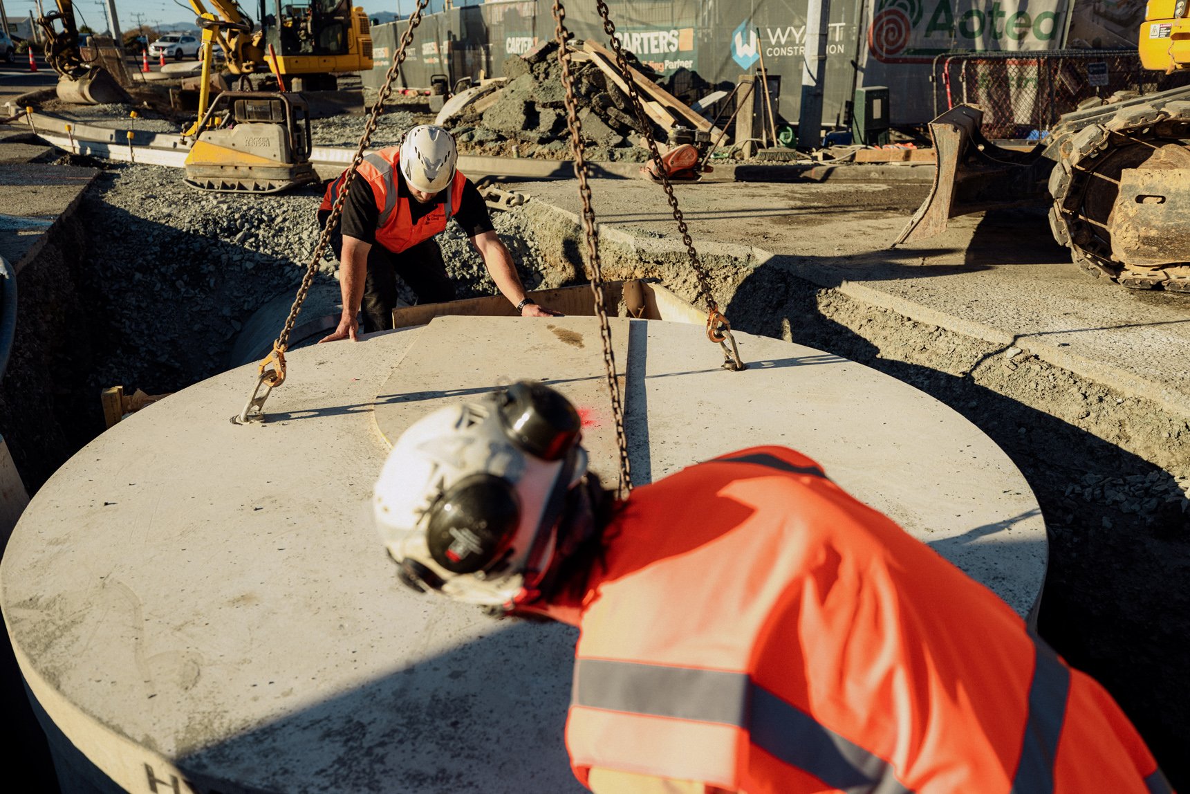 Construction workers wearing safety helmets and orange vests installing a large concrete pipe at a construction site, surrounded by construction equipment and piles of dirt.