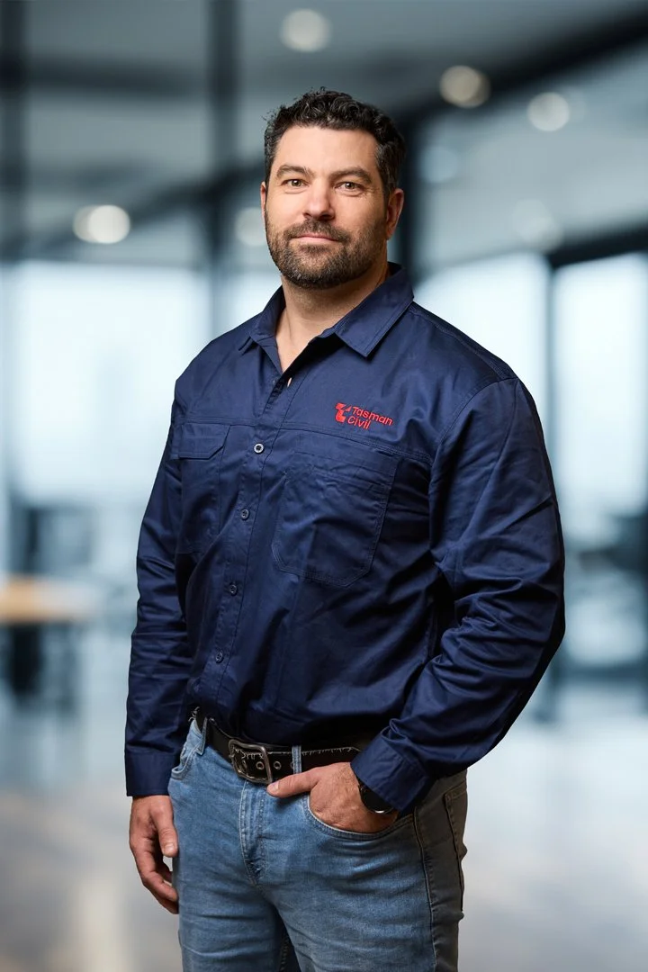 A man wearing a navy blueTasman Civil work shirt stands inside a modern office building with large windows.