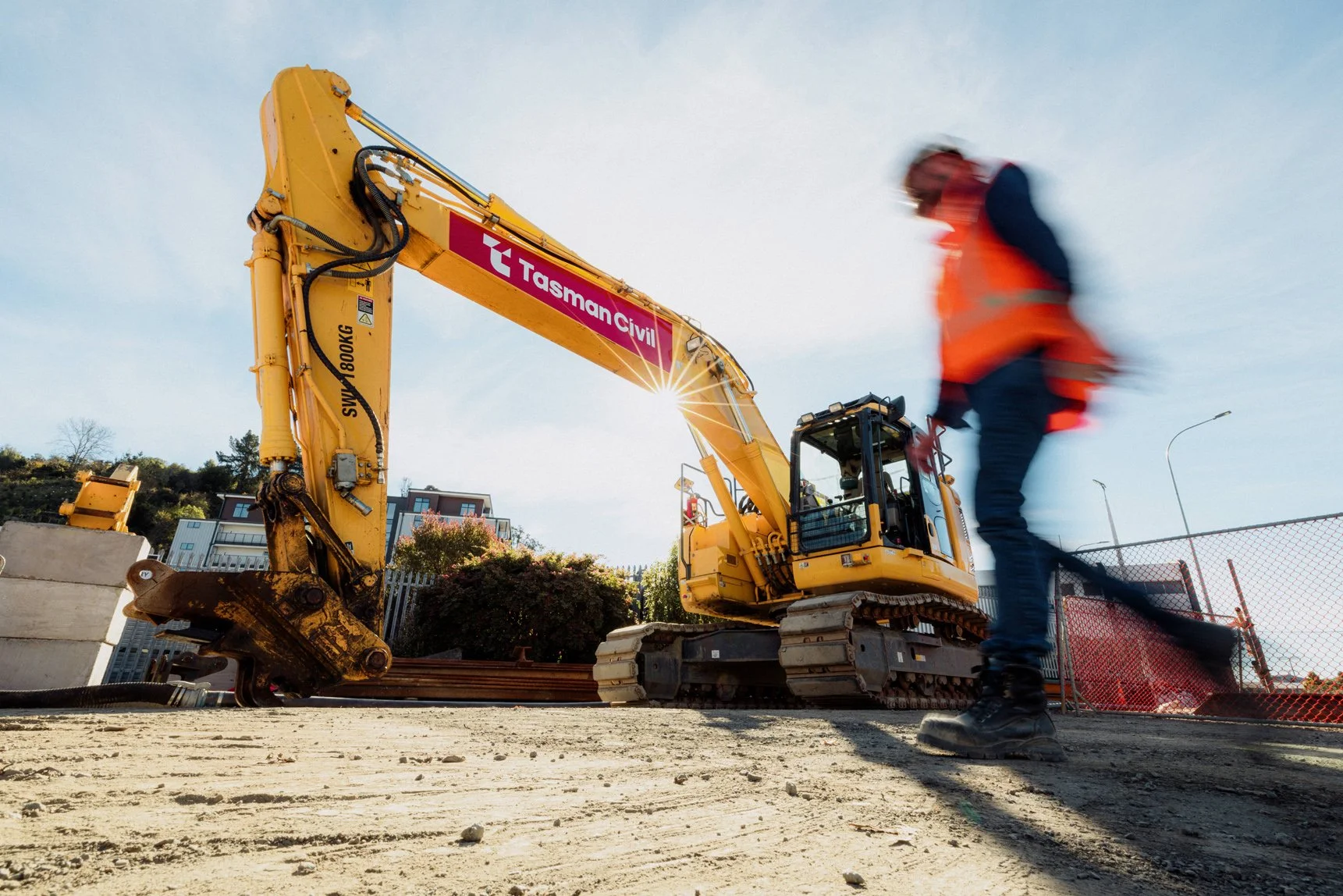 A yellow excavator with a red and white logo reading 'Tasman Civil' on its arm is operating at a construction site. A worker in an orange safety vest and dark clothing is walking past the machinery. The sky is clear and the ground is sandy with some concrete barriers and safety fencing nearby.