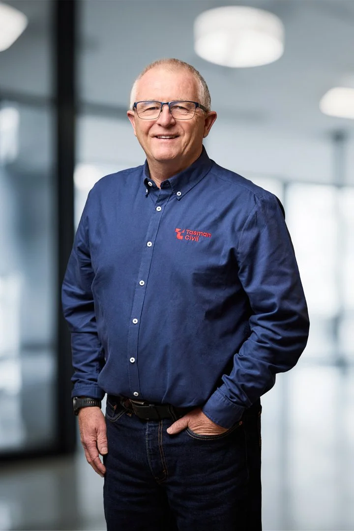A smiling man wearing glasses and a blue button-up shirt with the logo 'Tasman Civil' standing in a modern office environment.