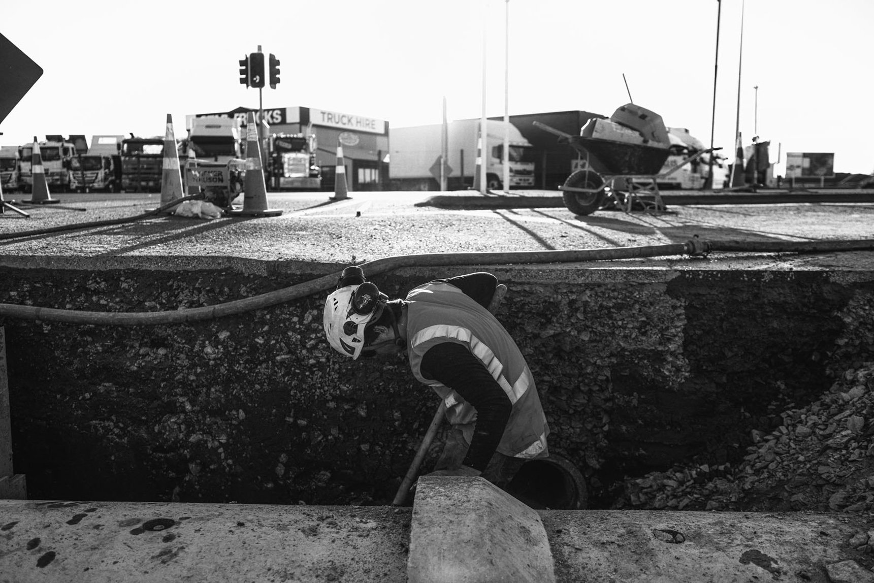 A construction worker wearing a safety vest and helmet is working on an underground pipe at a construction site. The scene shows traffic cones, a wheelbarrow, and construction equipment in the background.