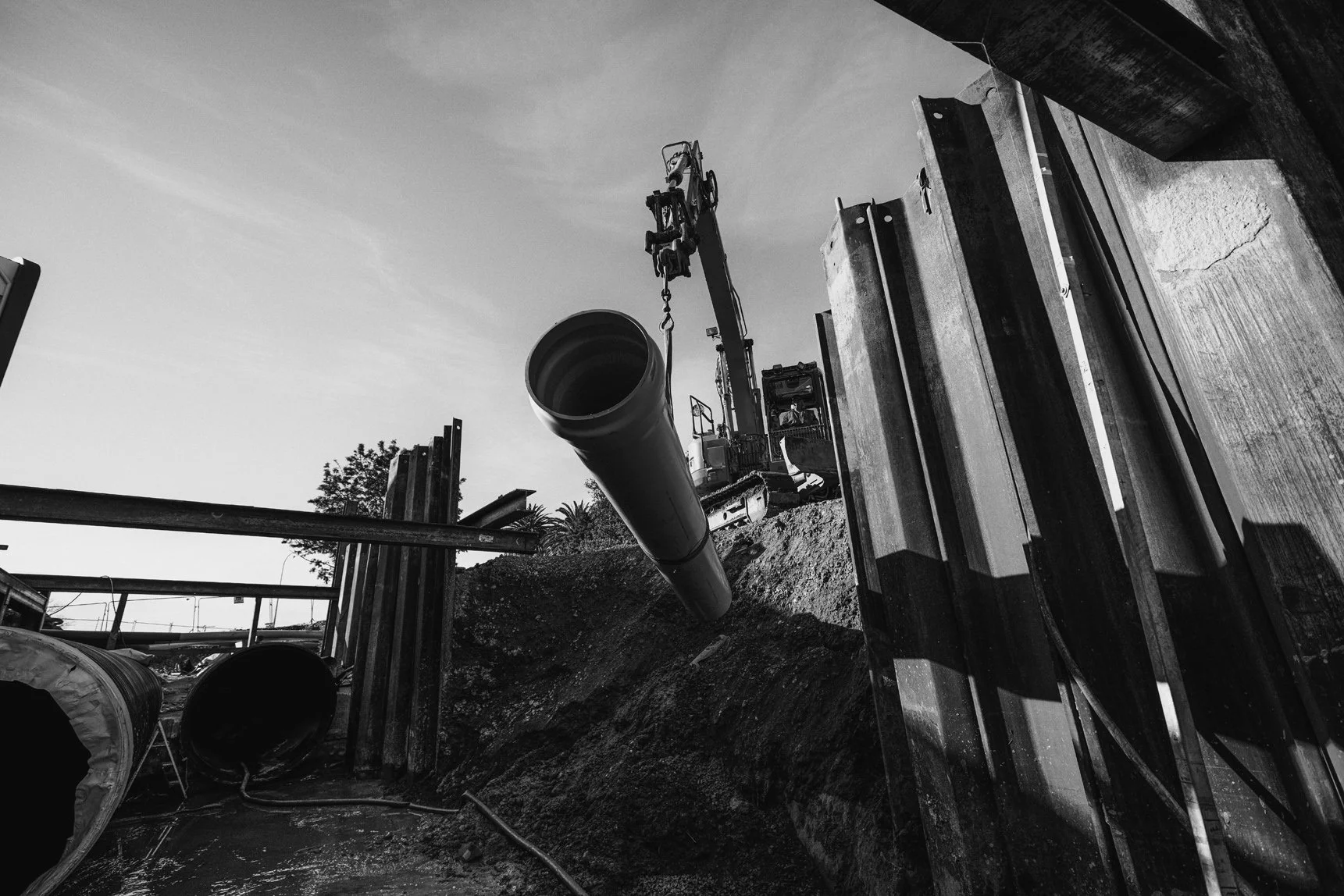 Construction site with a crane lifting a large pipe, dirt and construction materials on the ground, and trees in the background