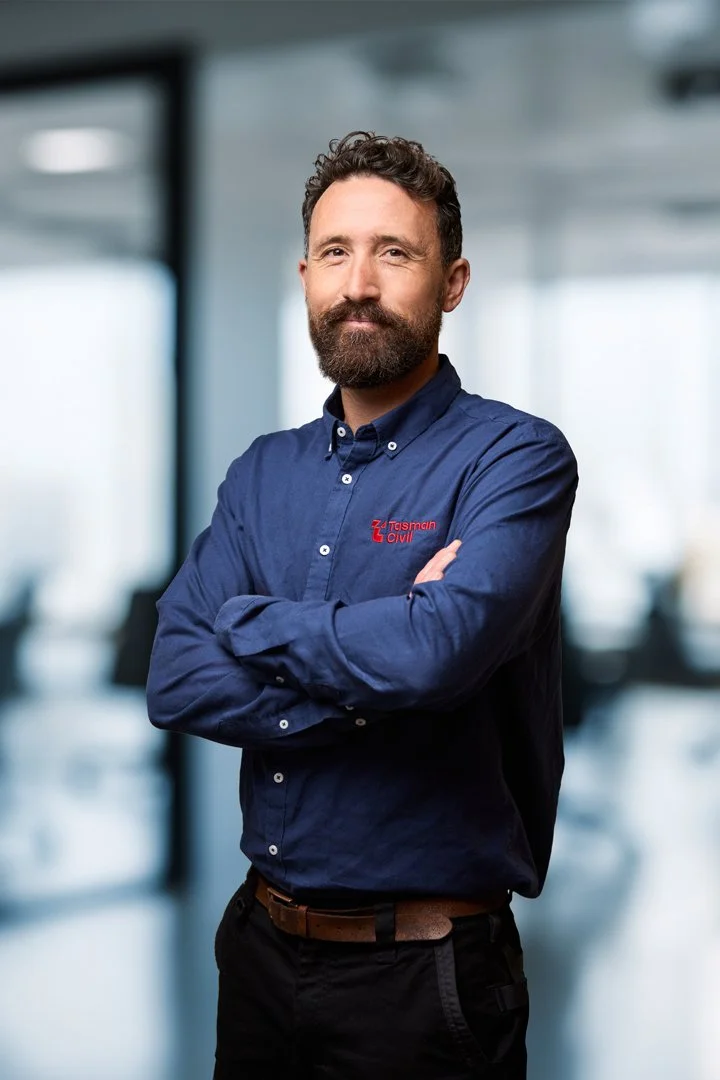 Man with a beard and mustache in a blue shirt with red text, arms crossed, standing in a modern office environment.