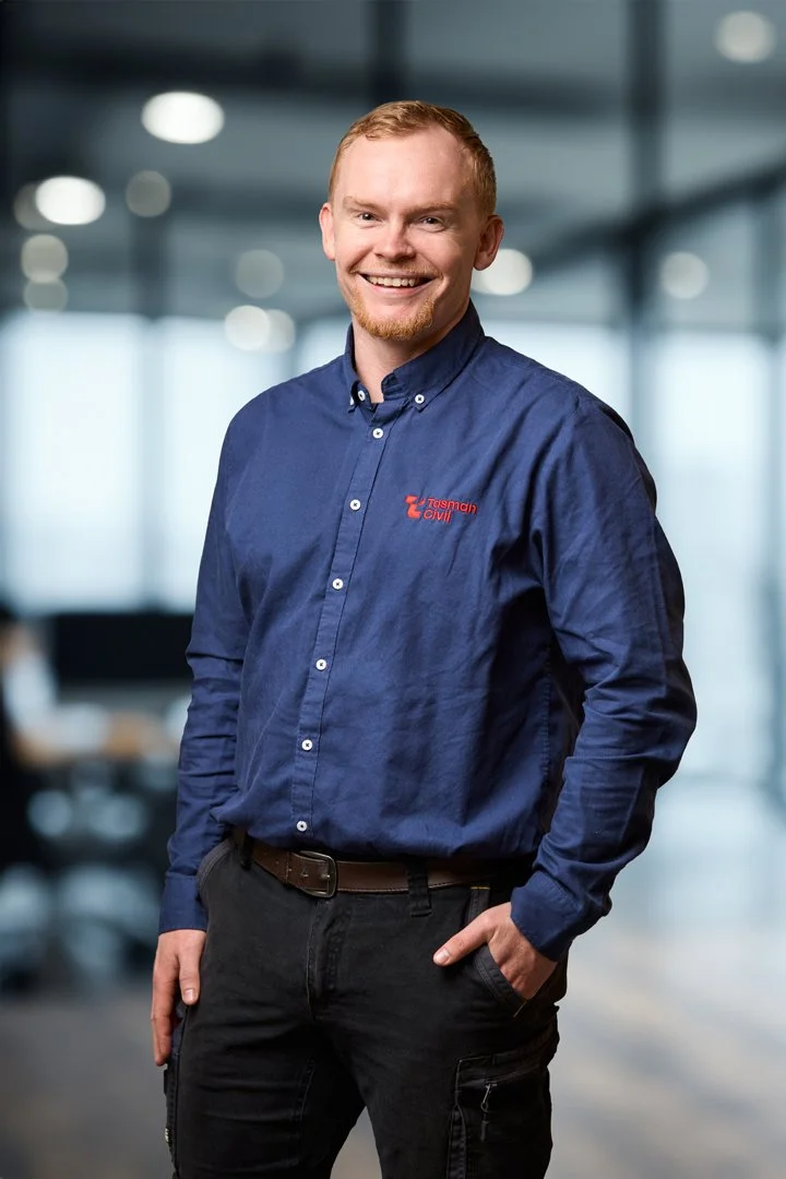 A young man with red hair and a beard, wearing a blue button-up shirt with a logo that says 'T elsewhere civil,' stands indoors in what appears to be an office environment, smiling confidently.