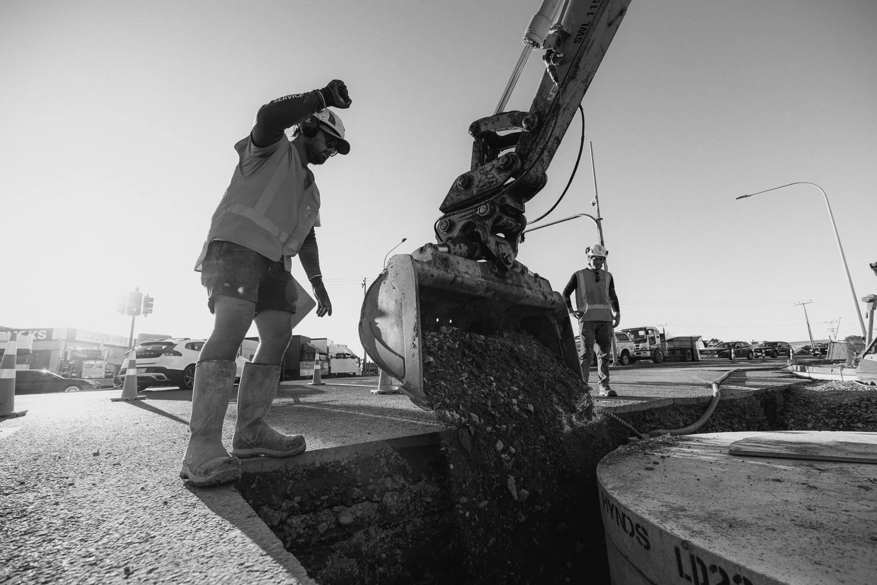 Construction workers operating an excavator to dig a trench on a paved street, with parked cars and traffic lights in the background during daylight.