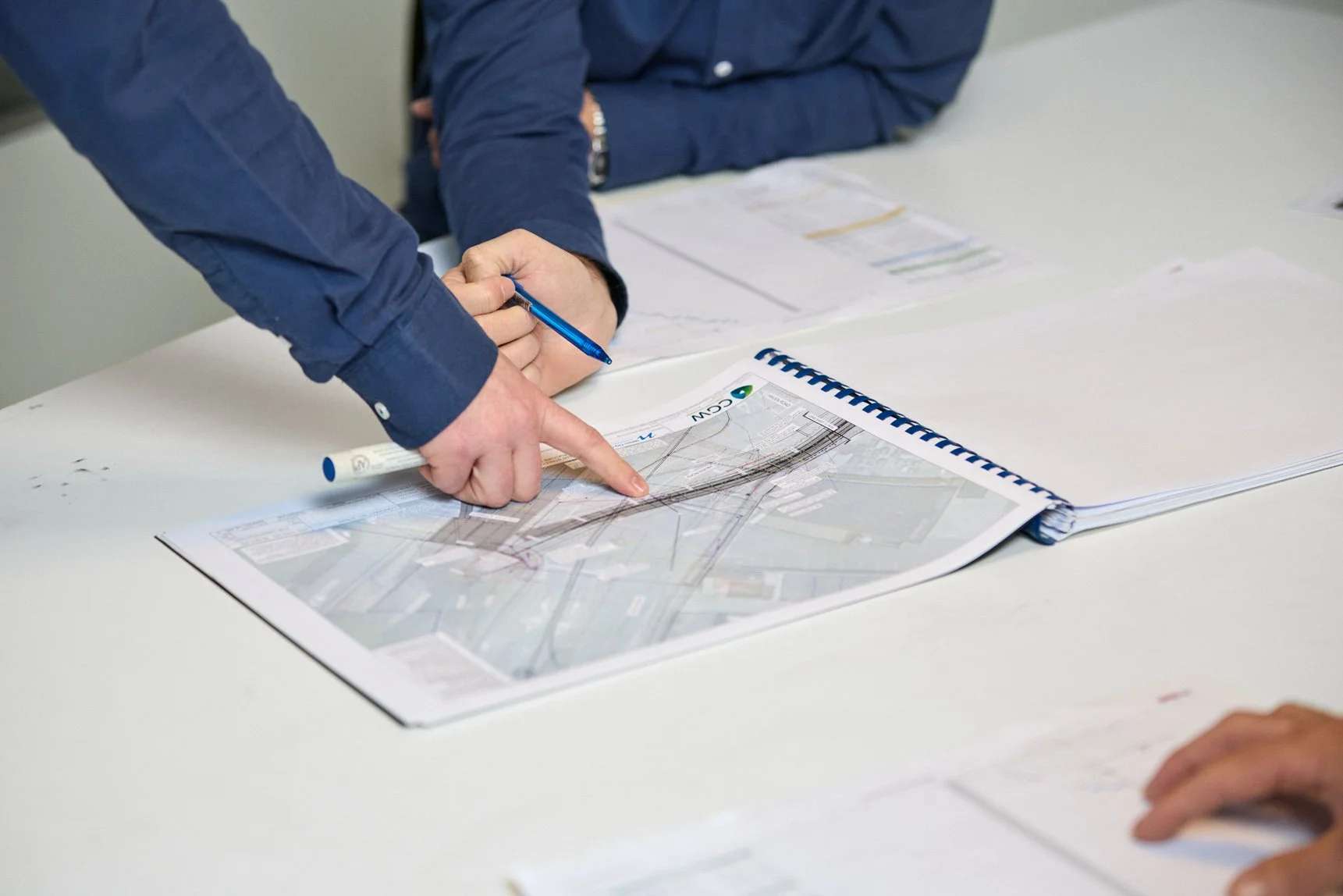Two people in blue shirts discussing a detailed map on a white table, with one person pointing at the map and the other holding a blue pen.