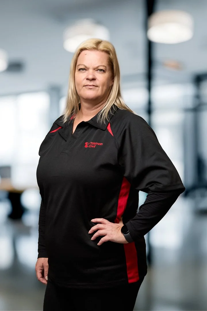 A confident woman with blonde hair wearing a black and red Tasman Civil polo shirt, standing in a modern indoor space with bright lighting and blurred background.