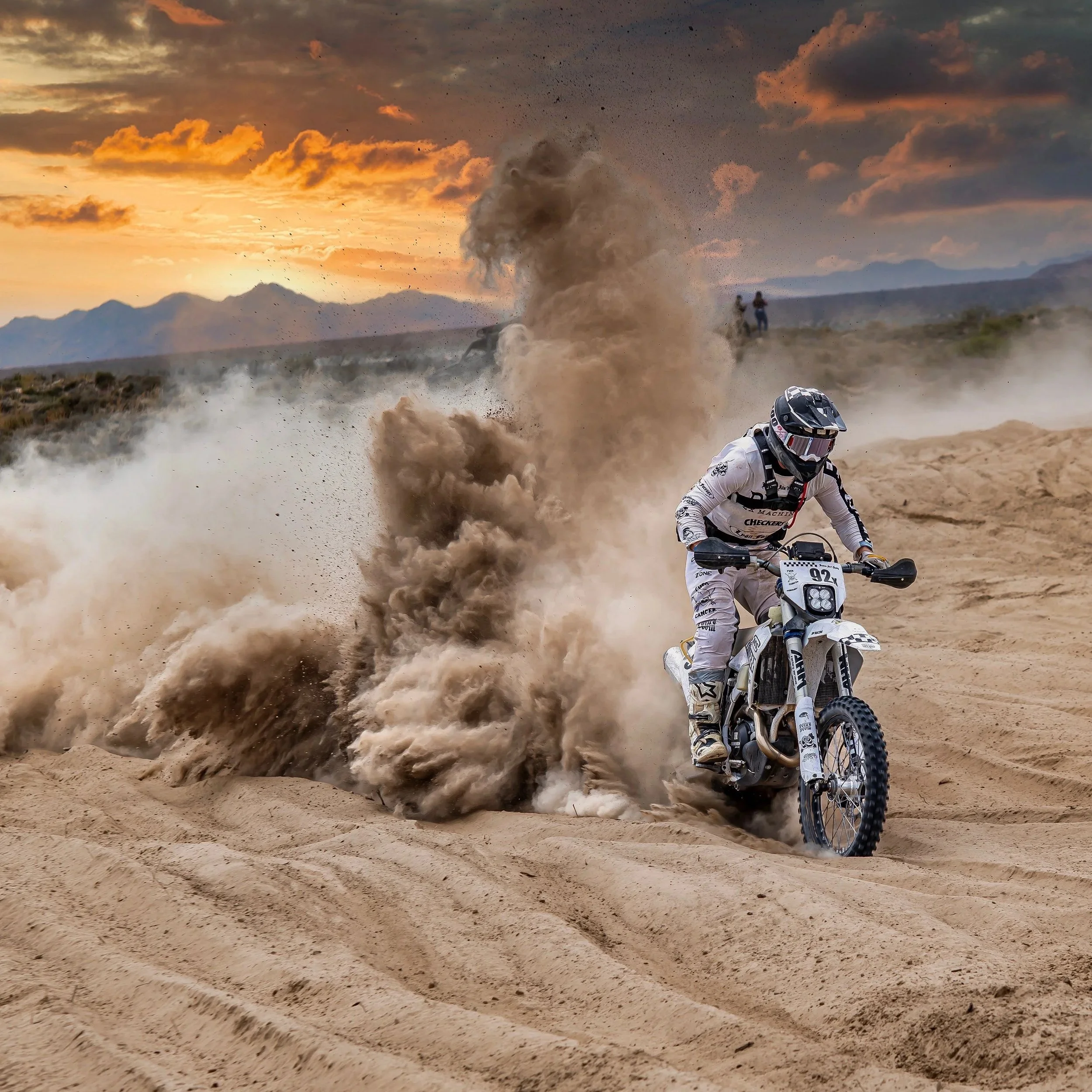 Racer Rescue - Motocross rider in white gear navigating sandy terrain at sunset, with dust and dirt flying, and mountains in the background.