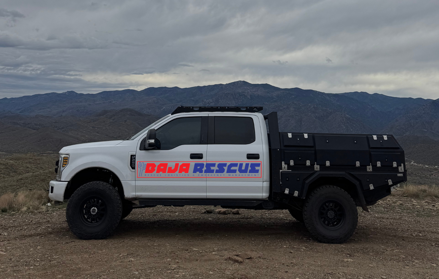 A white rescue truck with black cargo bed parked on rocky terrain with mountains and cloudy sky in the background.