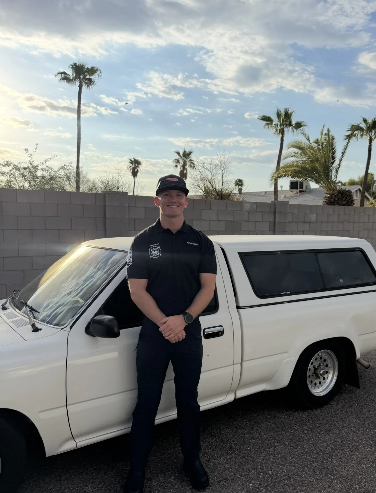 A young man in a police uniform standing in front of a white vehicle, with palm trees and a partly cloudy sky in the background.