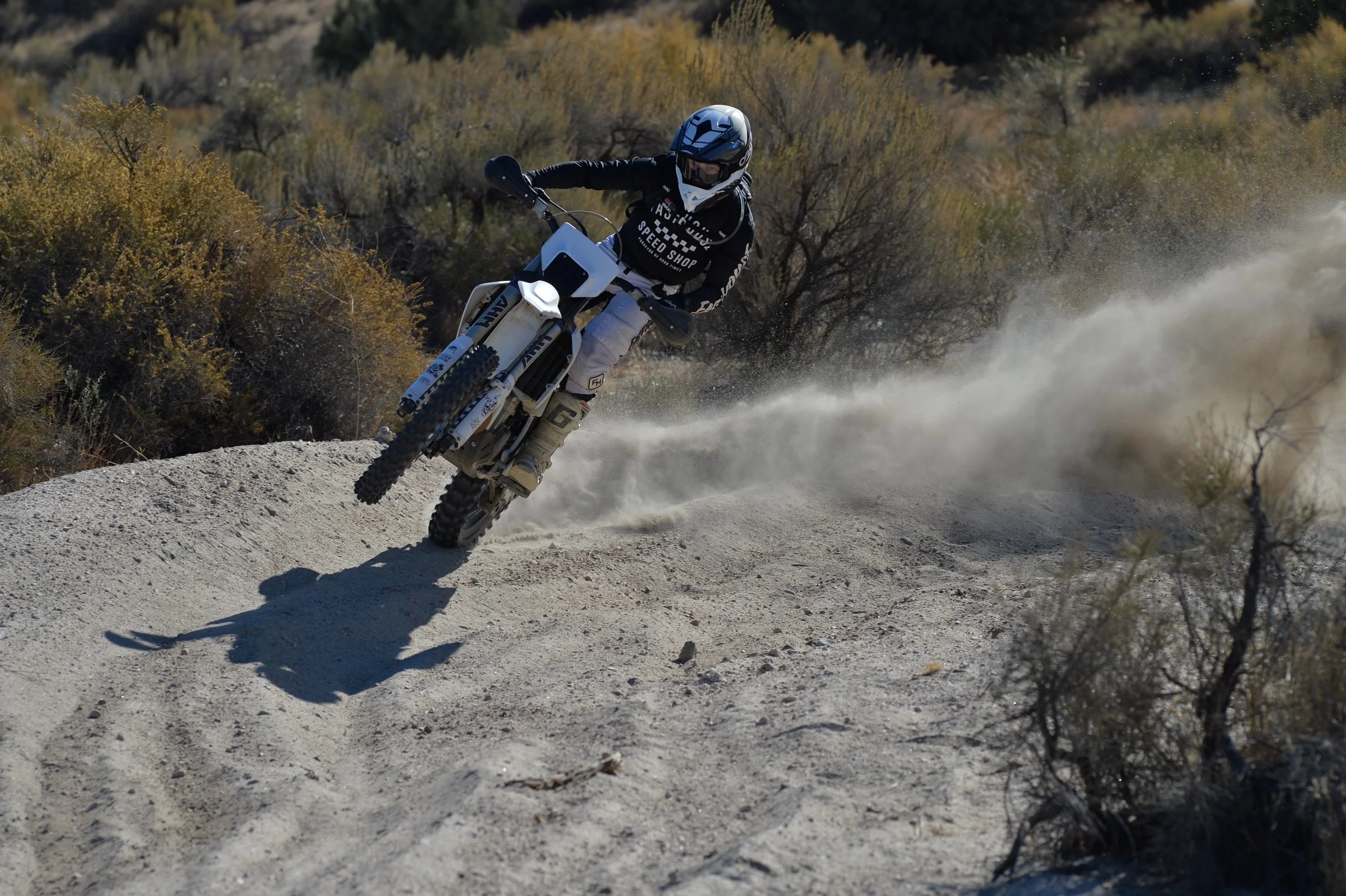 Motorcyclist riding off-road dirt bike on a dusty trail in a desert landscape.