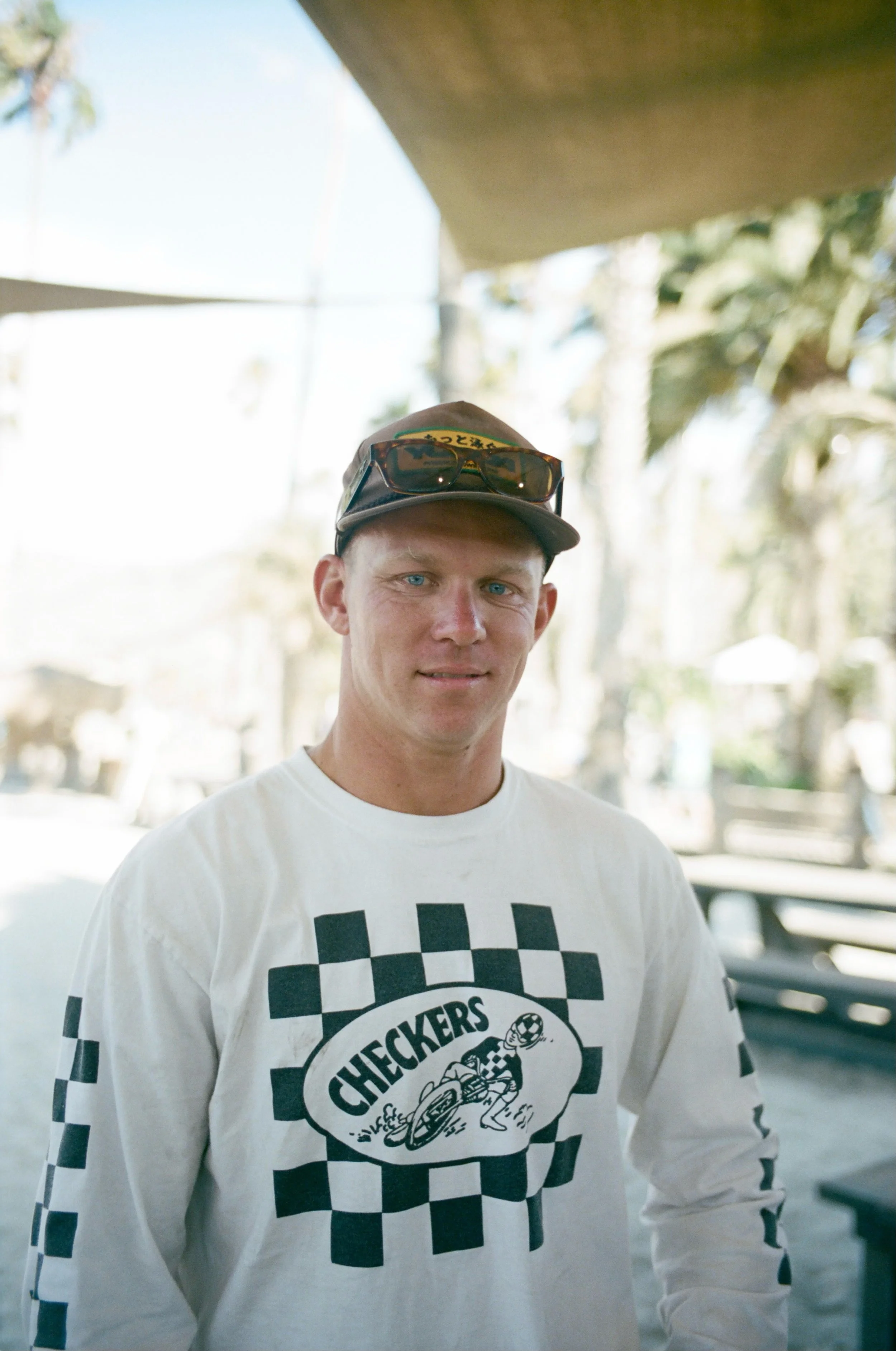 A young man with blue eyes and fair skin, wearing a white Checkers racing shirt, a brown cap, and sunglasses on his hat, standing outdoors with blurred trees and picnic tables in the background.