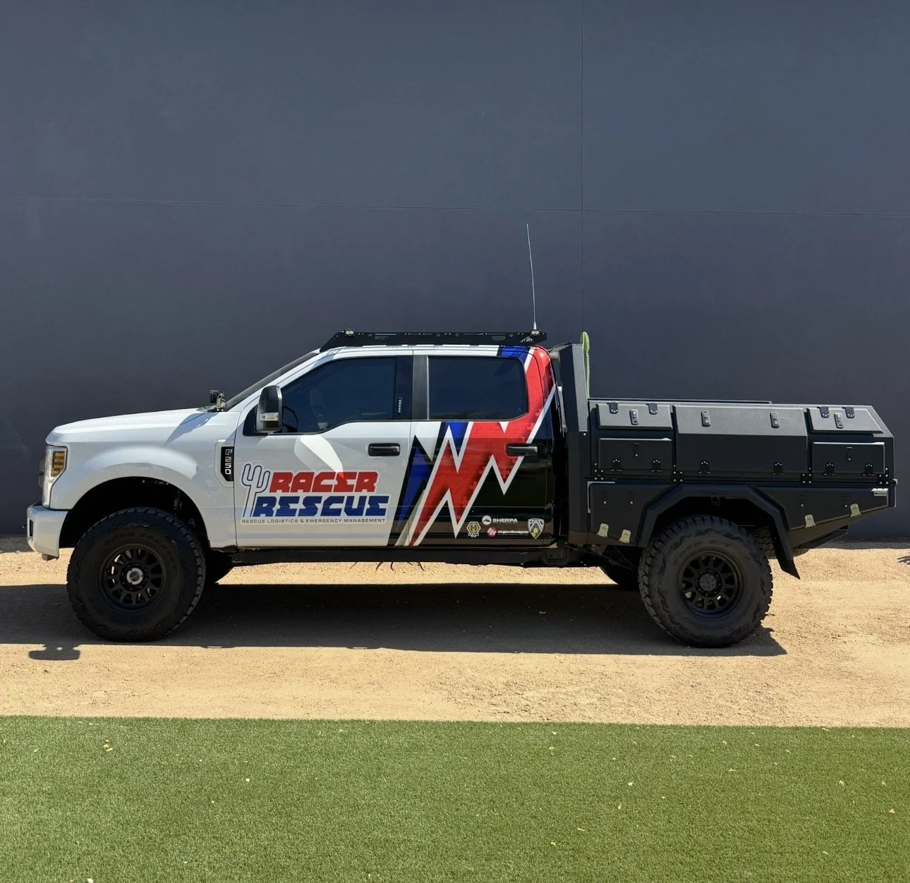 A white rescue truck with black cargo bed parked on a dirt road in a mountainous area under a cloudy sky, with a logo reading 'Baja Rescue' on the side.