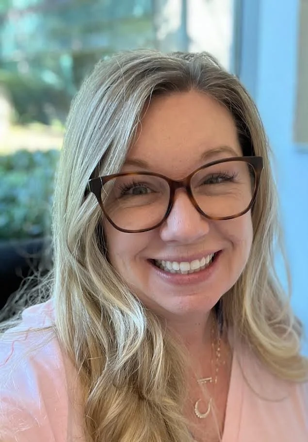 Close-up of a woman with blonde hair, wearing glasses with tortoiseshell frames, smiling indoors with a window and greenery in the background.