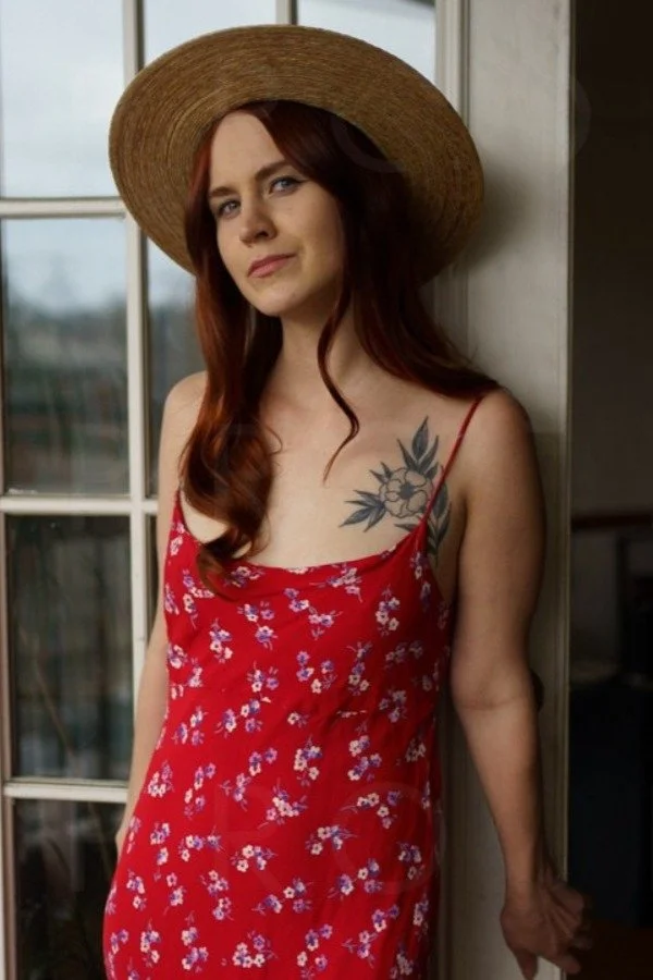 A red haired woman leaning against a glass paned door with a red patterned dress on and a straw hat.