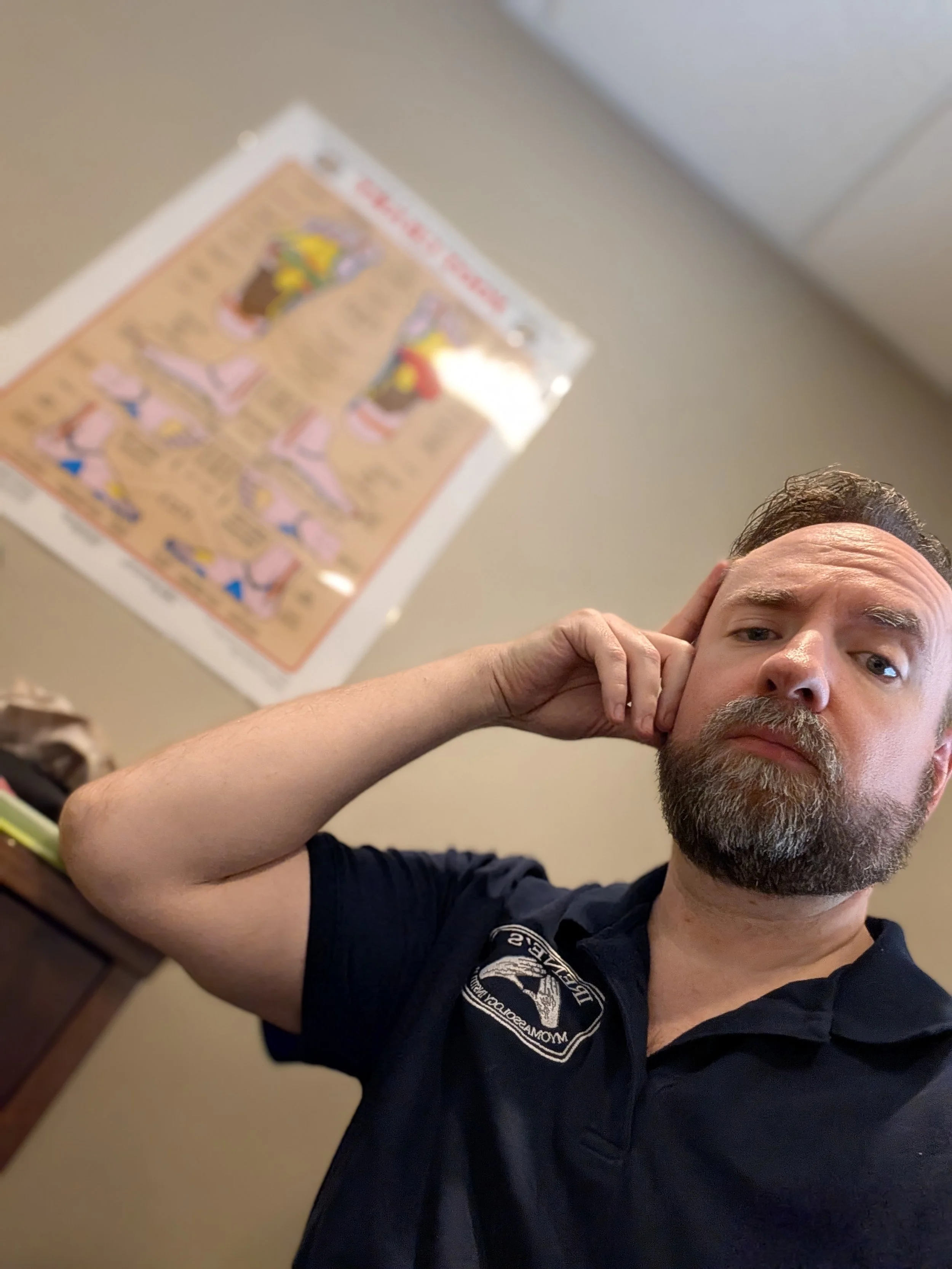 Man with beard and short hair holding his hand to his ear, wearing a black shirt with a circular logo, in a room with a beige wall and a colorful framed poster in the background.