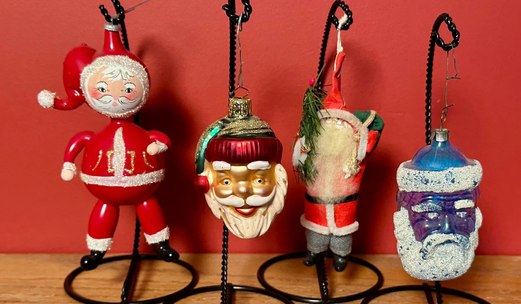 Four Christmas ornaments hanging on hooks against a red background. The first ornament is a Santa Claus figure with a round body, a Santa hat, and a jolly face. The second ornament is a Santa Claus face with a smiling expression, a red hat with a green brim, and a white beard. The third ornament is a Santa in a red and white suit with a small Christmas tree branch attached to his hat. The fourth ornament is a clear blue and purple snowman figure decorated with glitter.