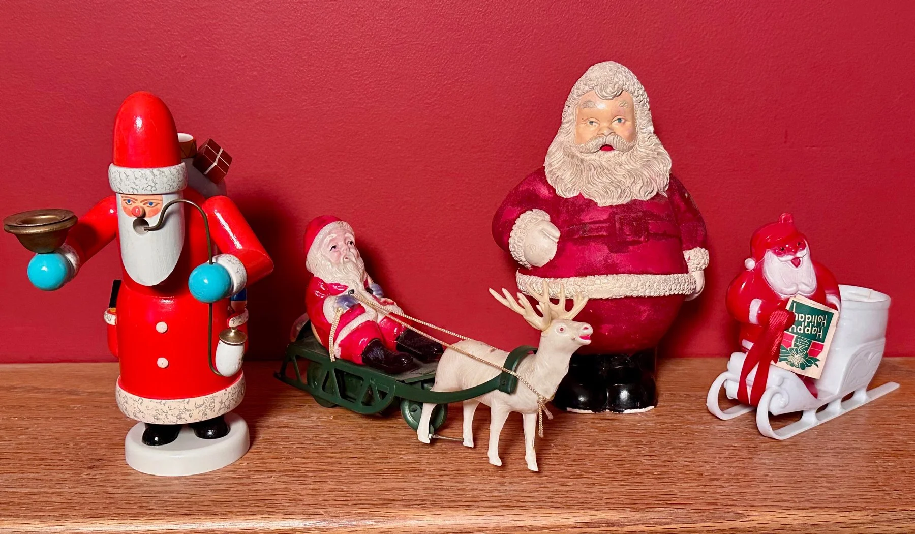 Collection of Christmas decorations on a wooden surface against a red wall. Includes a red and white Santa figurine with a pipe, a small Santa figure riding a sleigh with a reindeer, a Santa bust, and a small Santa in a sleigh with a Happy Holiday card.