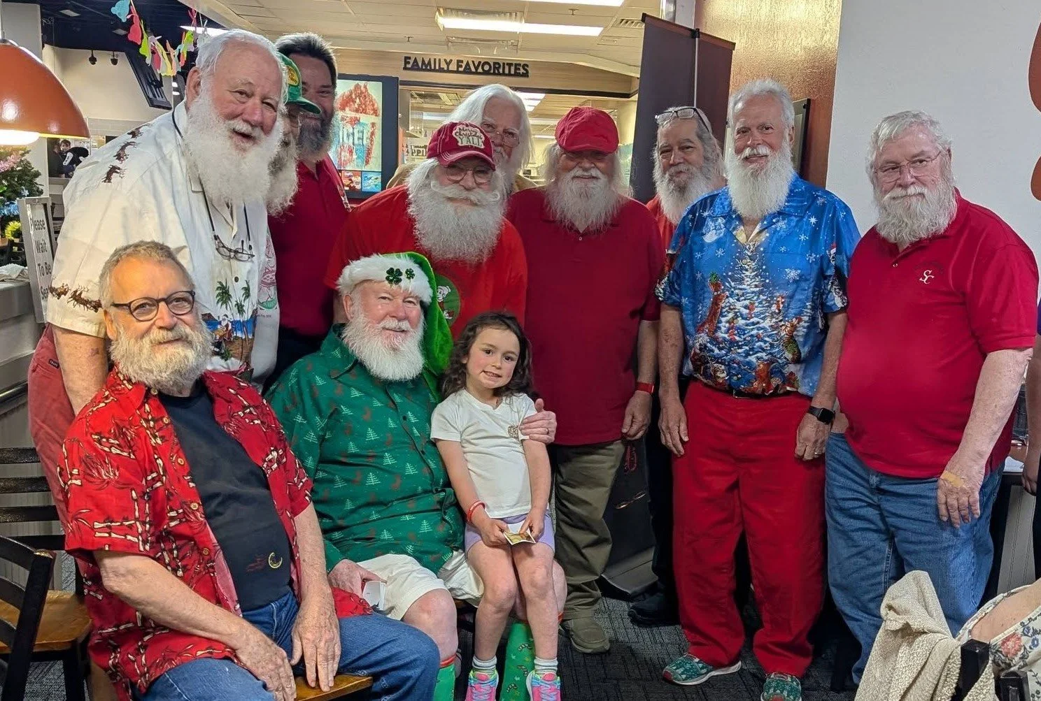 Group of men with white beards and a young girl posing for a photo indoors, some dressed in festive Hawaiian shirts, with a sign that says 'Family Favorites' in the background.