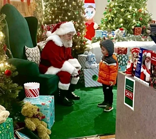 A young boy in a grey and orange jacket talking to Santa Claus in a store decorated for Christmas, with several decorated Christmas trees and wrapped gifts around.