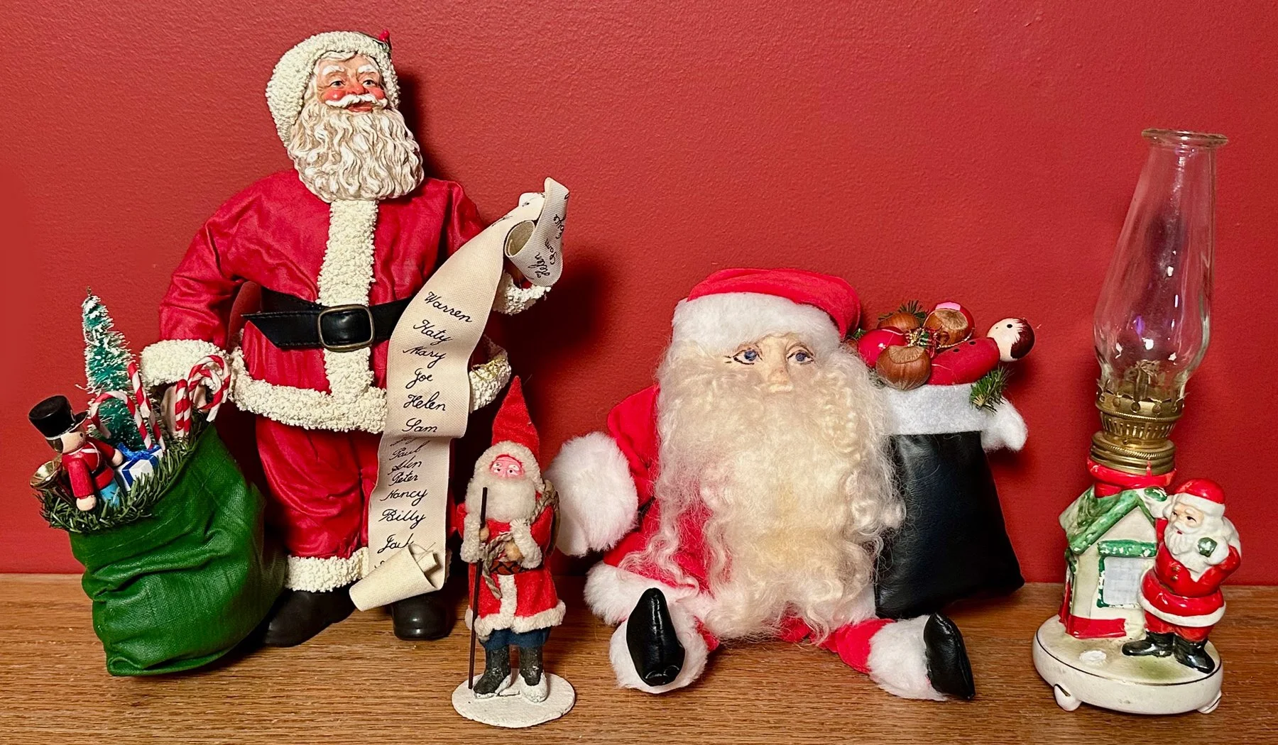 Christmas display with Santa figurine holding a list, a Santa plush doll, a small nutcracker, a small Santa figurine, a Santa sleigh with ornaments, a Santa candle holder, and a vintage oil lamp, all set against a red wall.