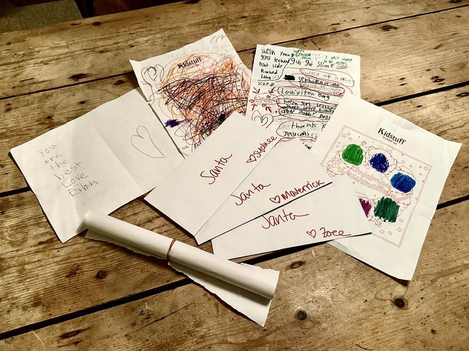 Collection of handmade and children's drawings, cards, and notes on a rustic wooden table, with colorful scribbles, hearts, and handwriting addressed to Santa.