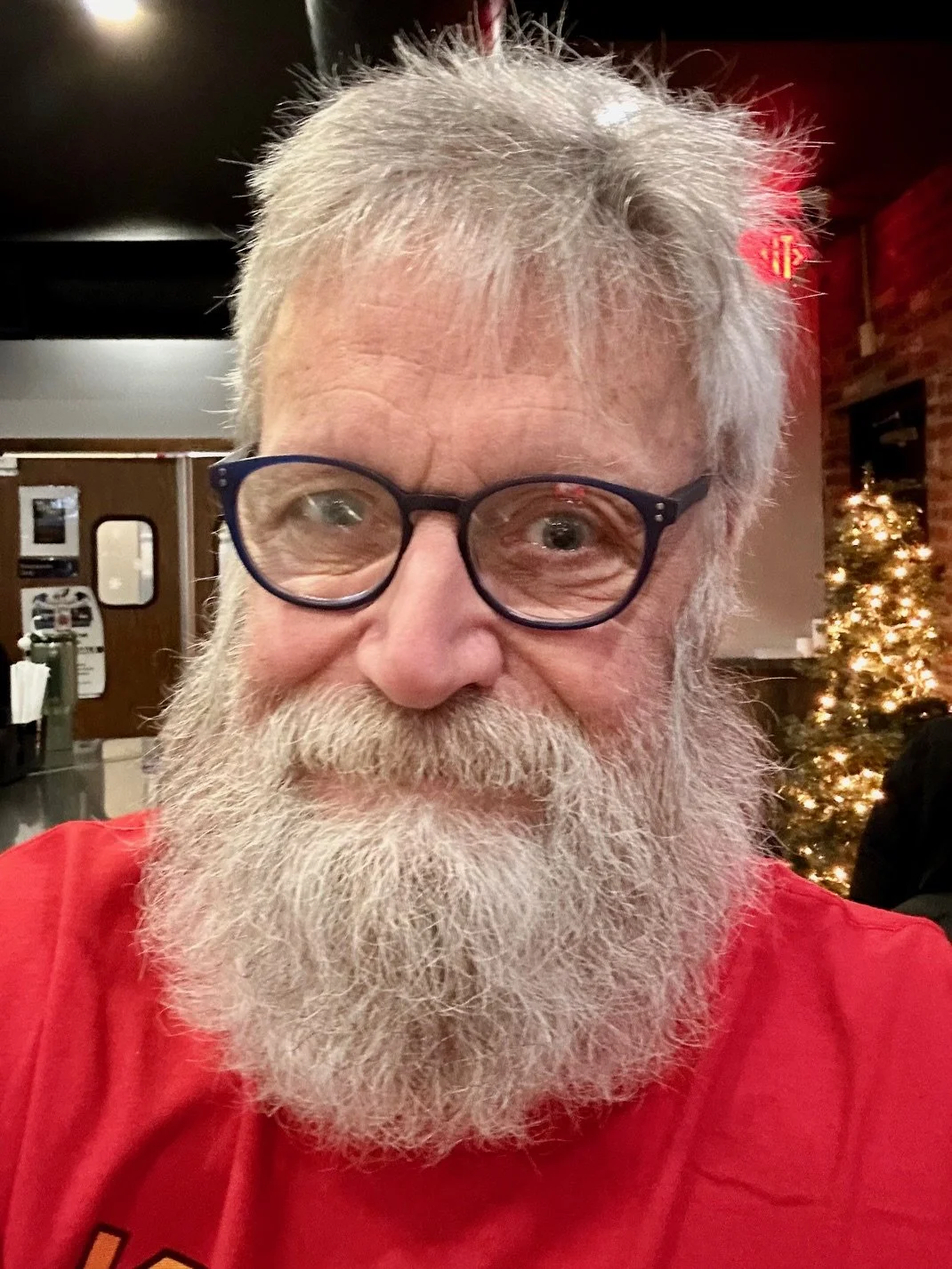 Close-up of an elderly man with white hair and beard, wearing dark glasses and a red shirt, indoors with Christmas tree and holiday decorations in the background.