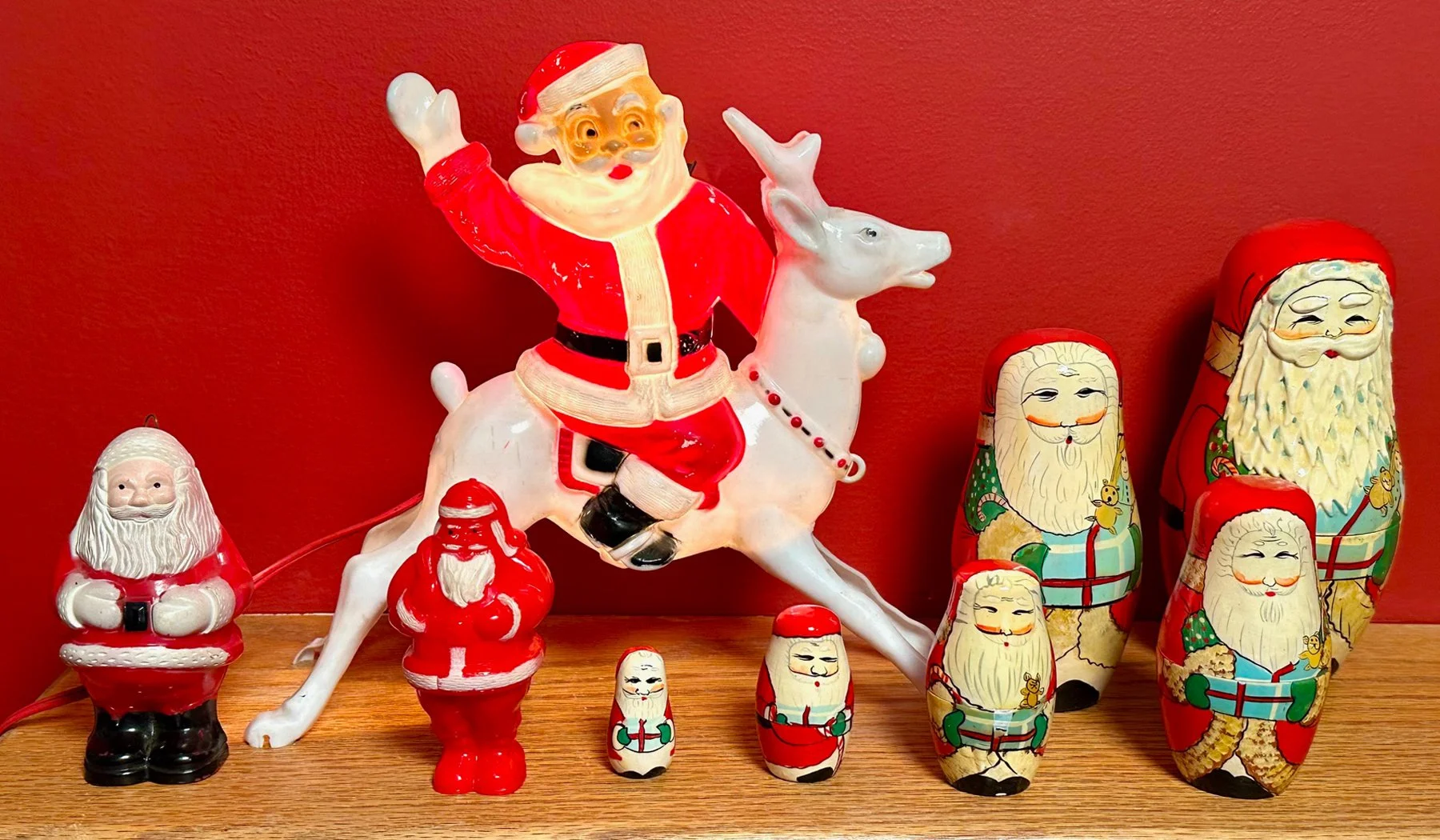 Collection of vintage Santa Claus decorations including a large Santa on a reindeer, several smaller Santa figurines, and Matryoshka dolls with Santa faces on a wooden surface against a red wall.