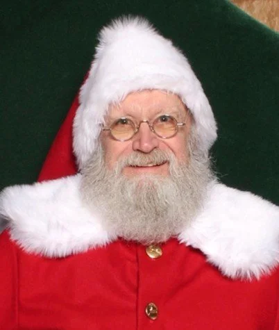 Man dressed as Santa Claus with a white beard, glasses, and a red suit with white fur trim, sitting in front of a dark green background.
