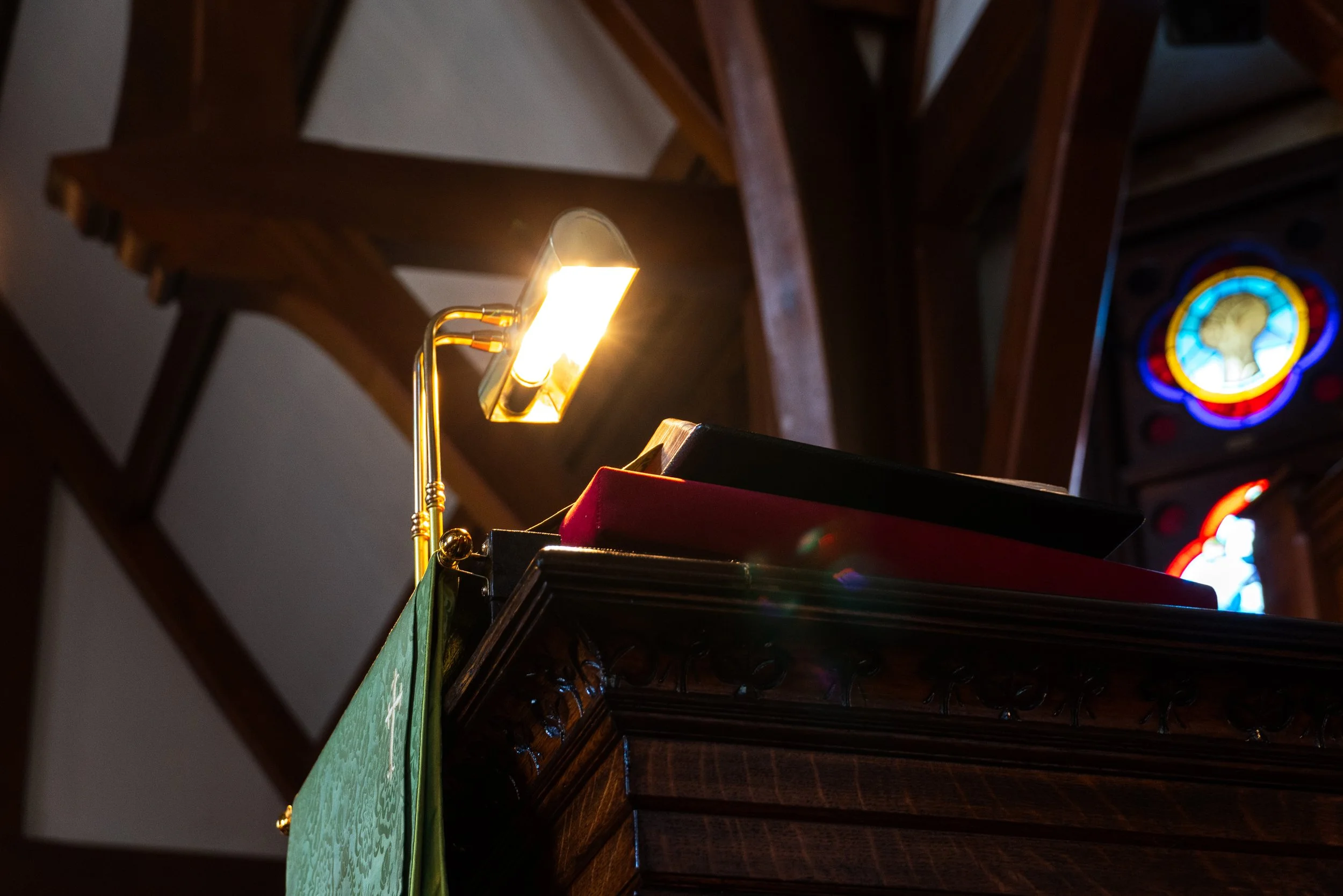 A close-up of a keyboard on a wooden pulpit with a reading lamp illuminating a book, in a church interior with stained glass window visible in the background.