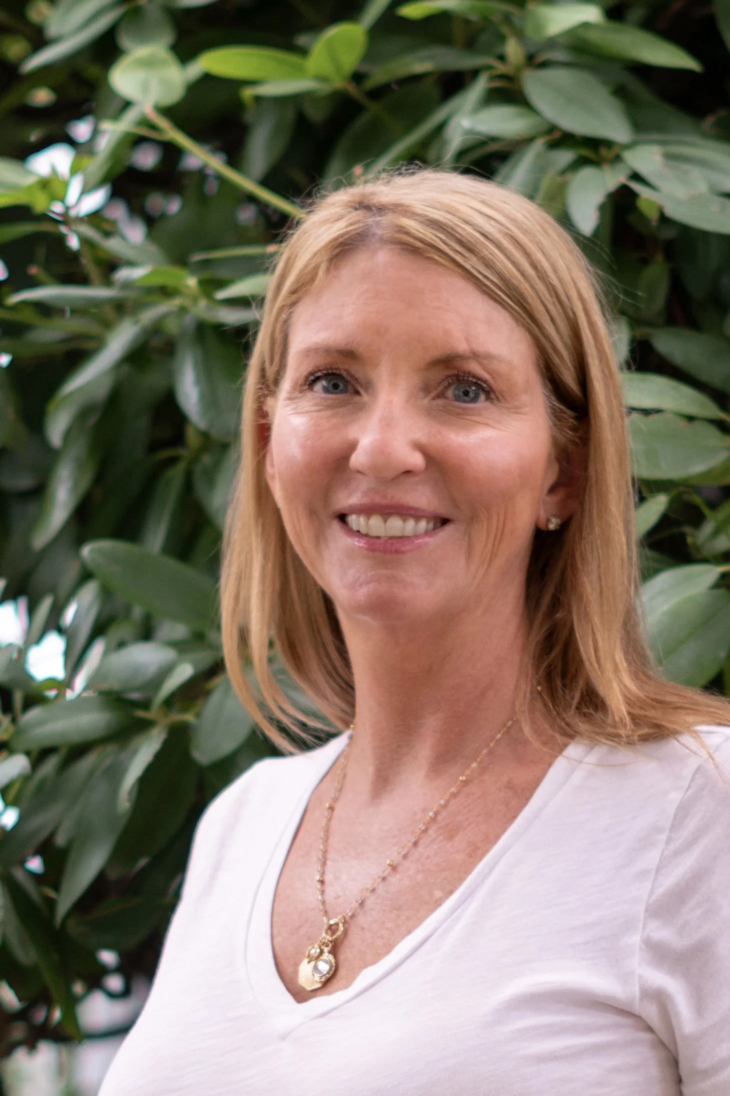 Close-up of a smiling woman with blonde hair, blue eyes, wearing a white V-neck top and gold jewelry, standing in front of leafy green plants.