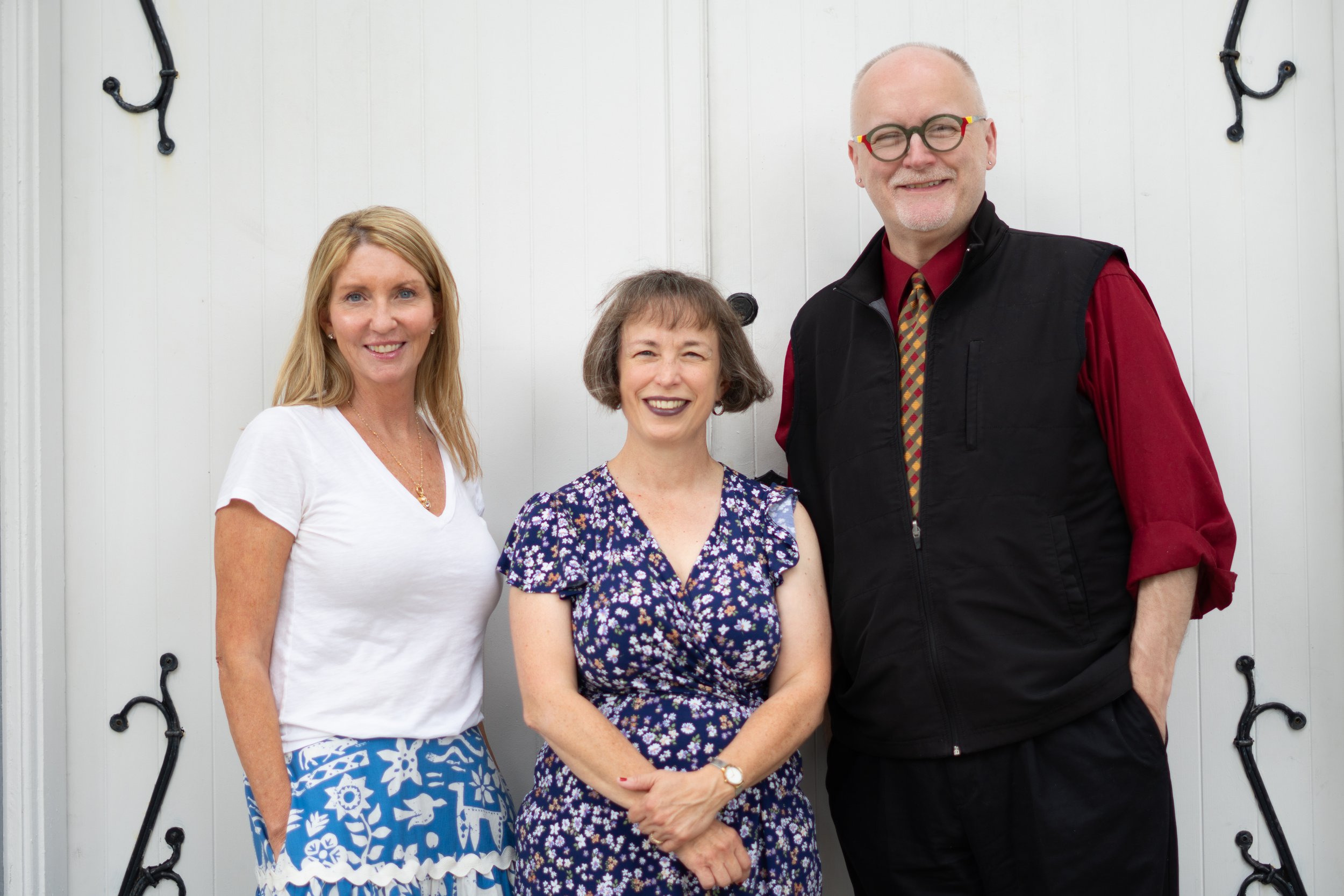 Three women and one man standing in front of a white door with black decorative hinges. The woman on the left has long blonde hair and is wearing a white top and blue patterned skirt. The woman in the middle has short brown hair and is wearing a floral dress. The man on the right has a bald head, glasses with red and yellow frames, and is dressed in a red shirt with a black vest and a patterned tie.
