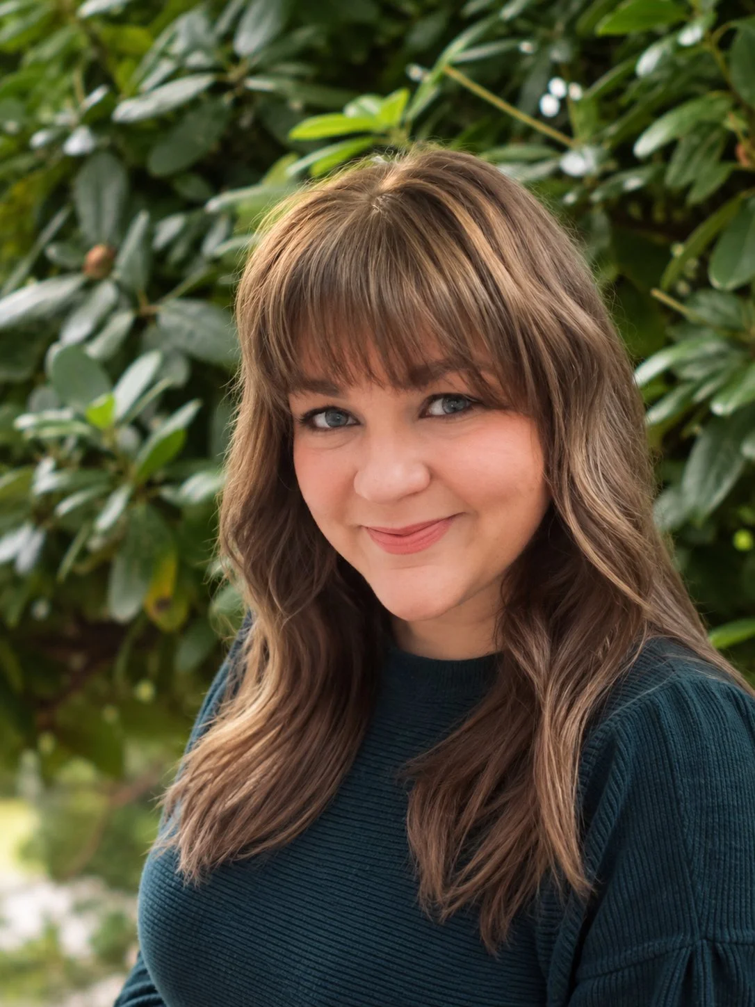 A woman with shoulder-length brown hair and bangs smiling outdoors with a bushy green background.