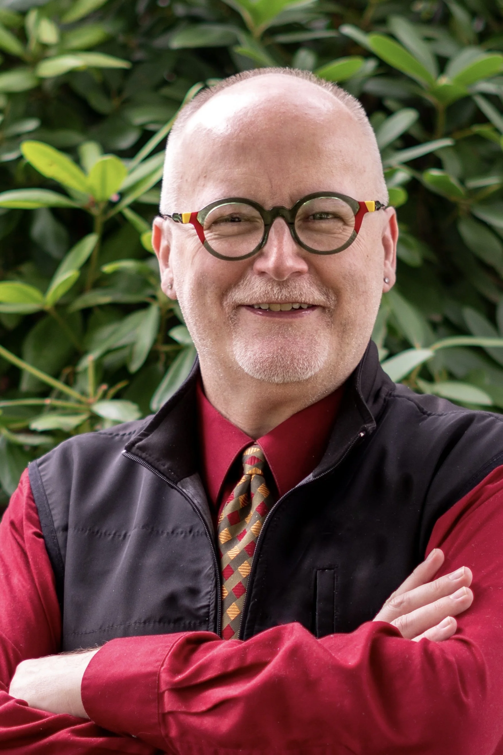 A man with a bald head and glasses smiling, wearing a black vest over a red shirt, with a patterned tie, in front of green foliage.