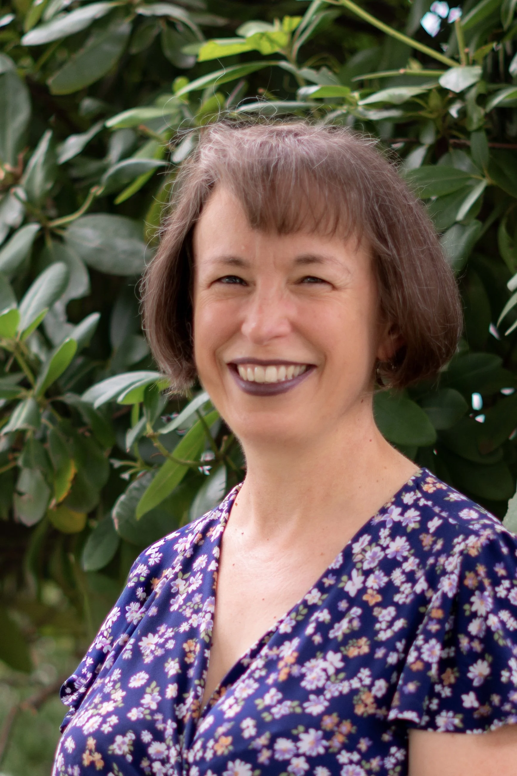 A woman with short brown hair and blue eyes smiling outdoors in front of green leafy bushes, wearing a navy blue dress with a pink and white floral pattern.