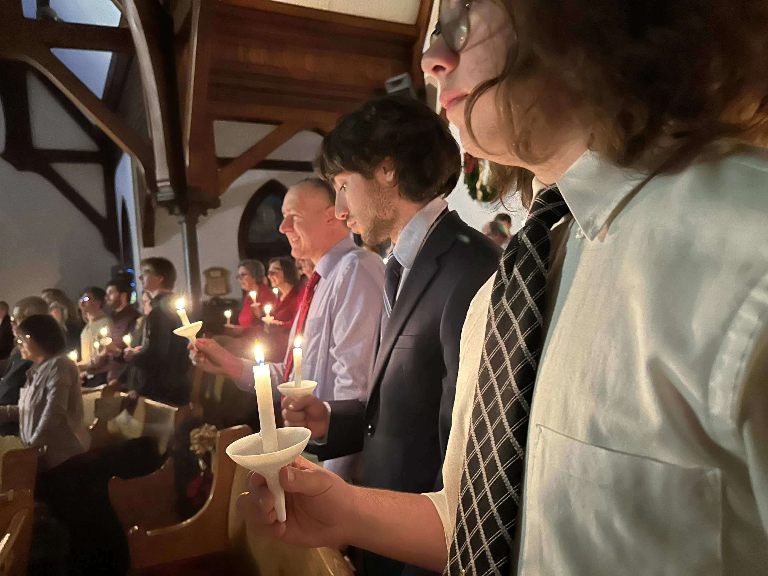 People participating in a candlelit ceremony, holding lit candles with white holders, in a room with wooden beam ceilings engaged in a solemn activity.